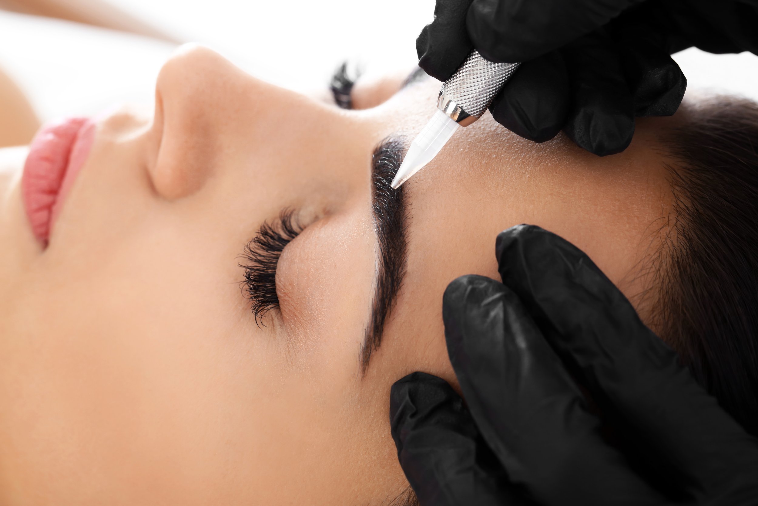 A woman receiving a cosmetic eyebrow tattooing or microblading procedure, lying down with her eyes closed, while a technician uses a tattoo pen with a needle on her eyebrows, wearing black gloves.