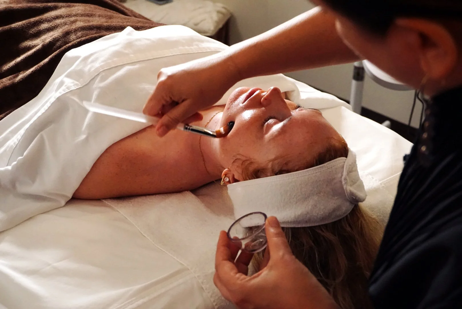 A person lying down on a spa bed receiving a facial treatment from an aesthetician using a brush and tools.