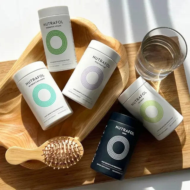 Five Nutrafol supplement bottles, a glass of water, and a wooden scalp massager on a wooden tray with sunlight.