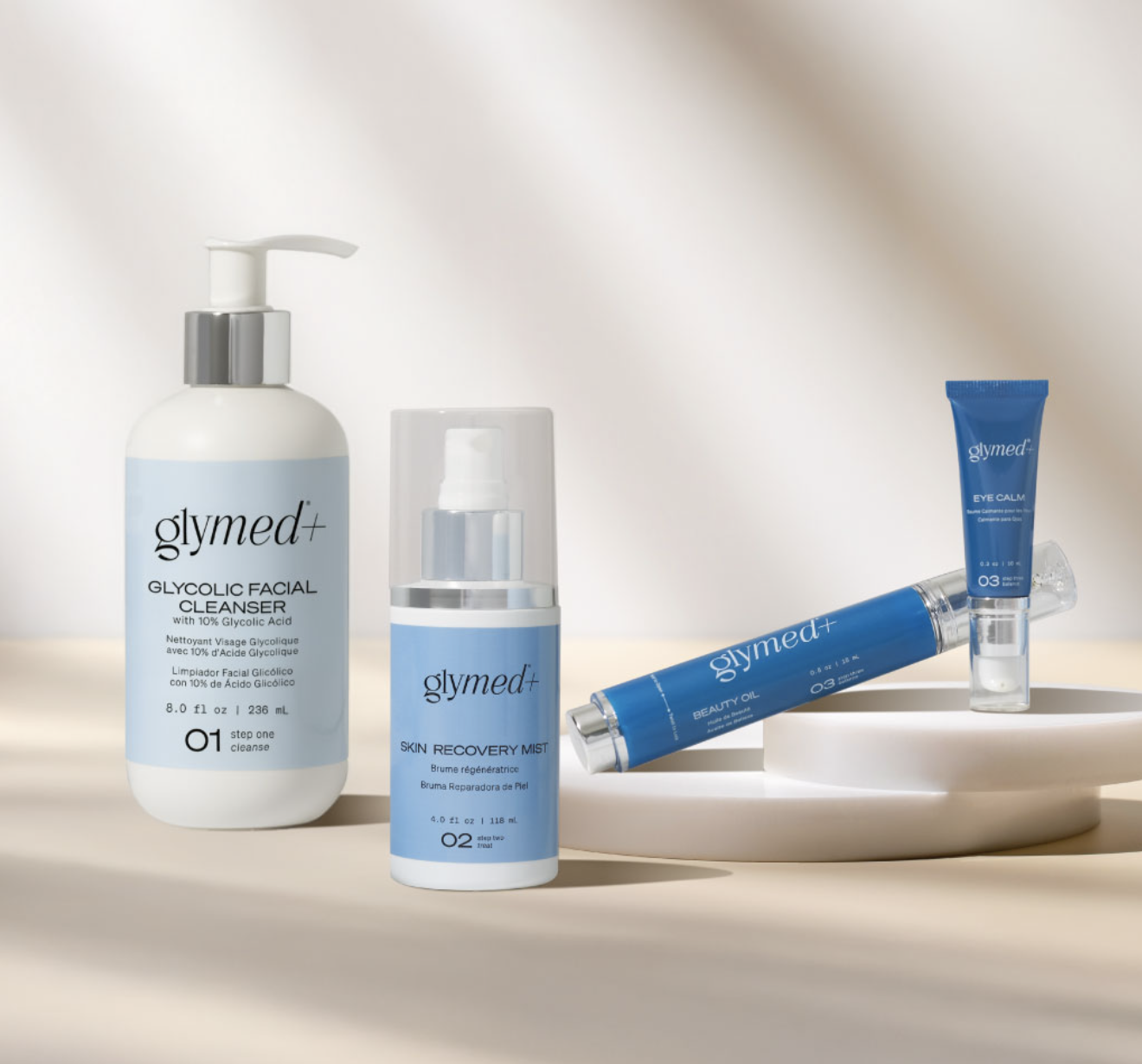 Skincare products including glycolic facial cleanser, skin recovery mist, beauty oil, and eye calm balm from the brand Glymed+ displayed on a white surface with a plain background.