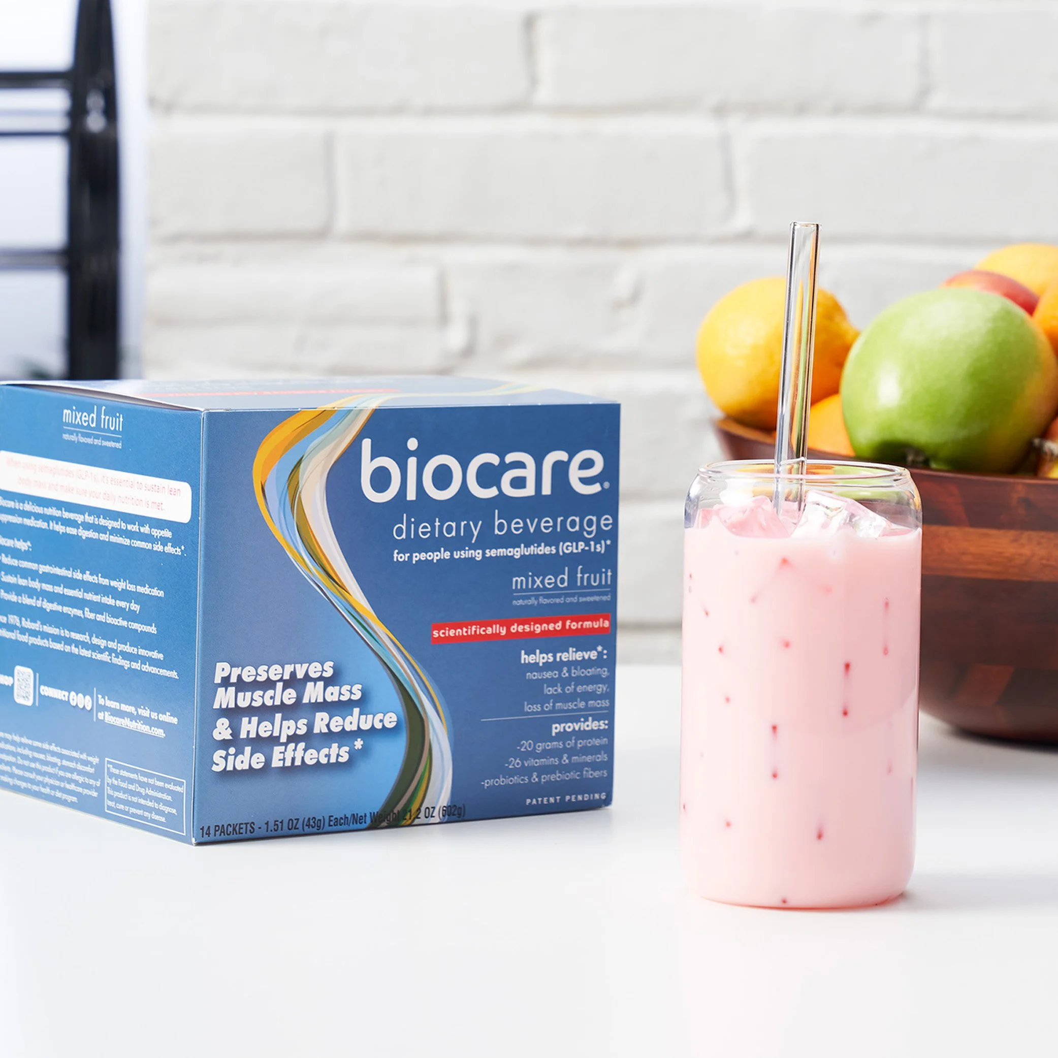 A box of Biocare mixed fruit dietary beverage packets, a glass of pink mixed fruit beverage with a straw, and a bowl of green and yellow apples on a white surface.