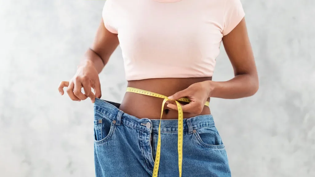 Person measuring waist with a tape measure, wearing oversized jeans and a pink crop top.