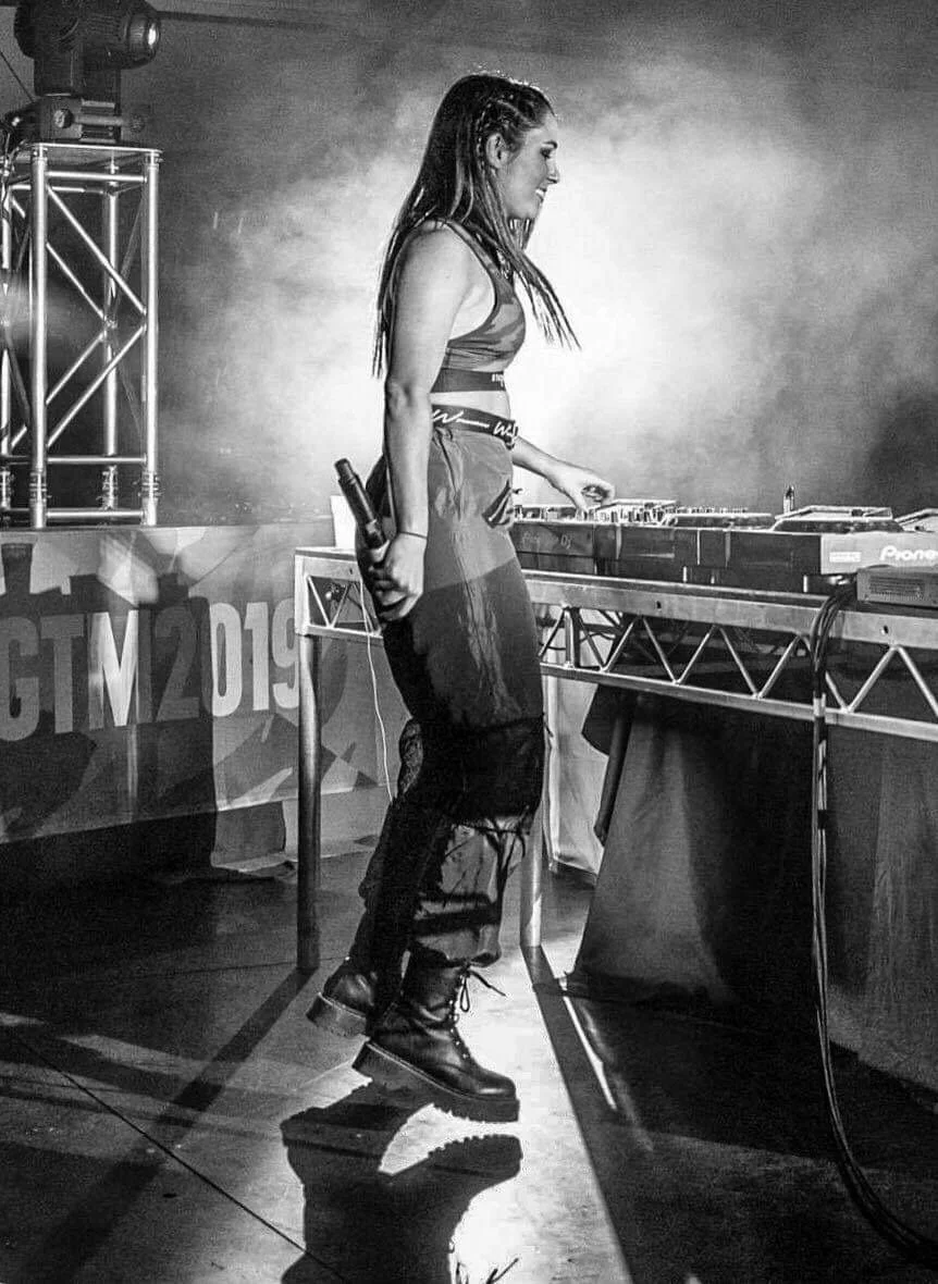 A female DJ in a sleeveless top and ripped jeans, holding a microphone, standing behind DJ equipment on stage at a concert or music event, with lighting and stage setup visible.