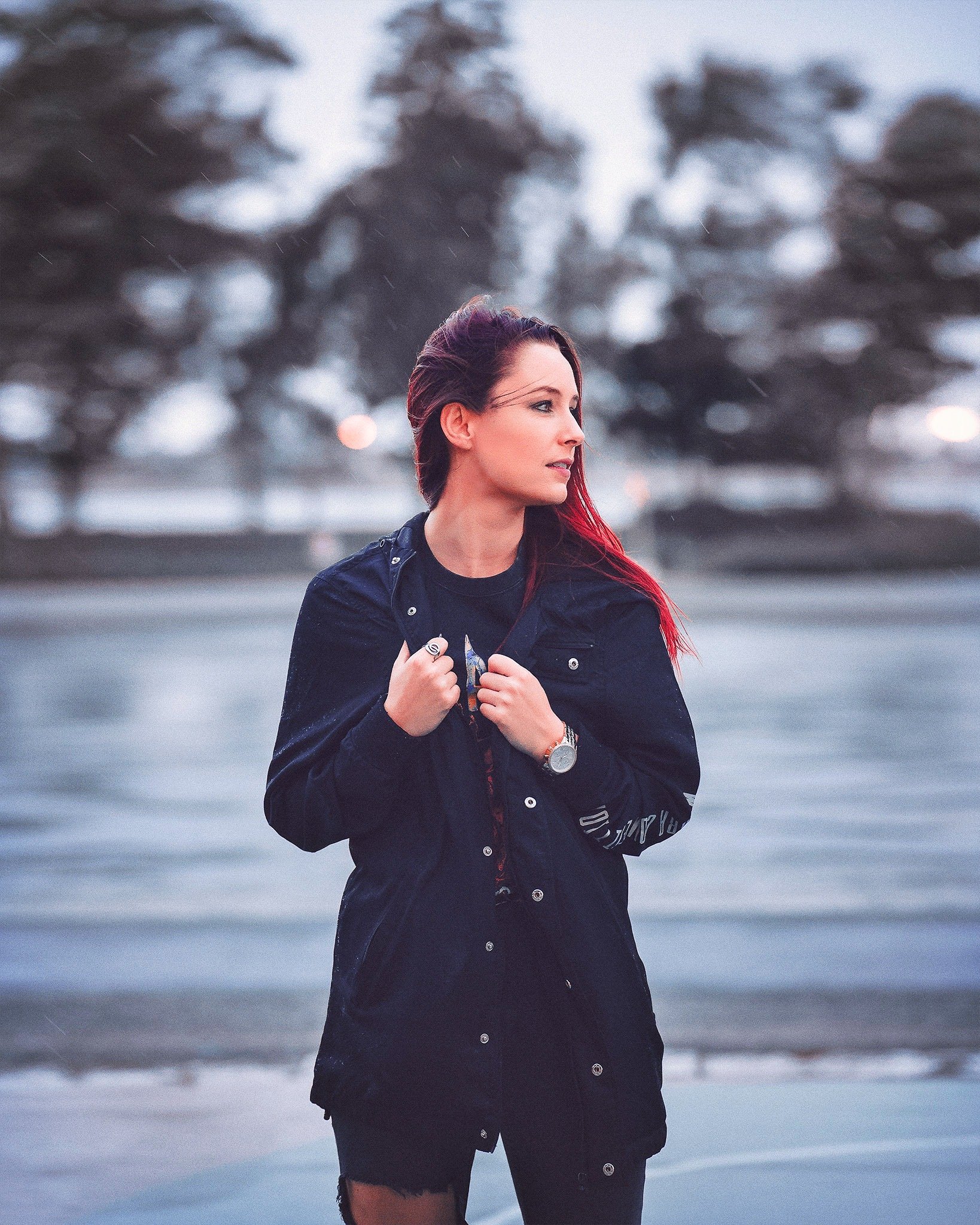 Young woman with red hair standing near water during sunset, wearing a black jacket and watch, with trees blurred in the background.