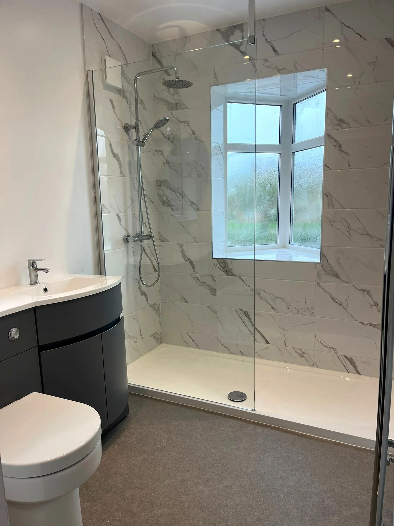 A modern bathroom with a walk-in shower featuring a large window and marble tiles, a small dark vanity with a sink, and a white toilet, all with a neutral color scheme.