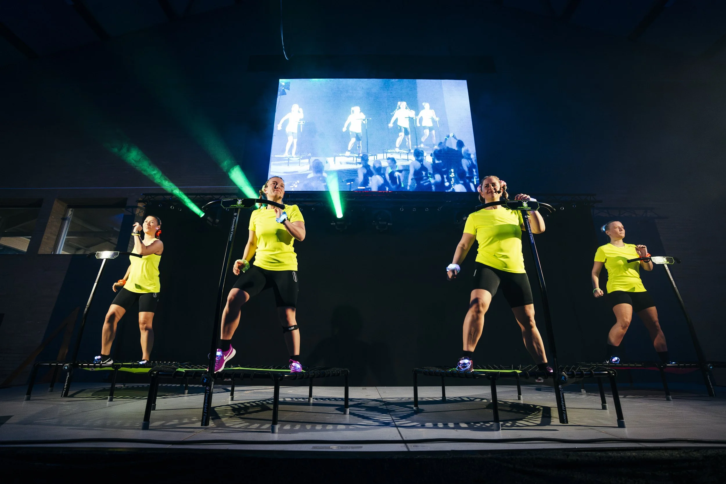 Kvinder deltager i jump fitness ved et event med stor skærm i baggrunden, der viser deres præstationer.