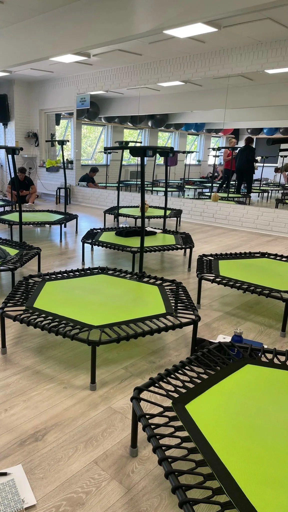 Et fitnesscenter med trampoliner med grønne overflader, væg med spejle og ansatte samt kunder, der forbereder sig på aktivitet.