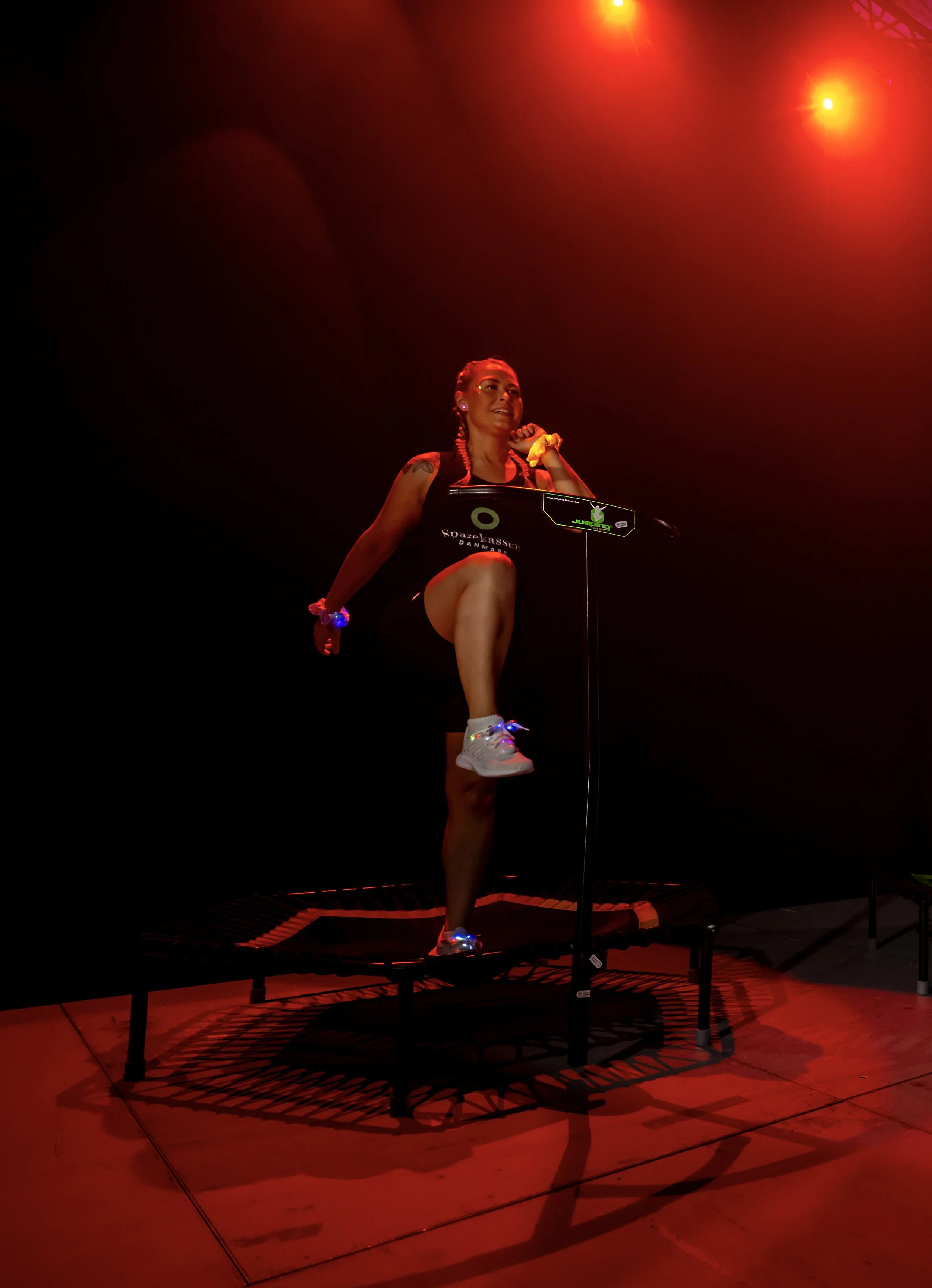 Instruktør Majbritt Jensen står på en jump fitness trampolin med et ben løftet, er klædt i sportsudstyr, og er under lys med rød belysning.