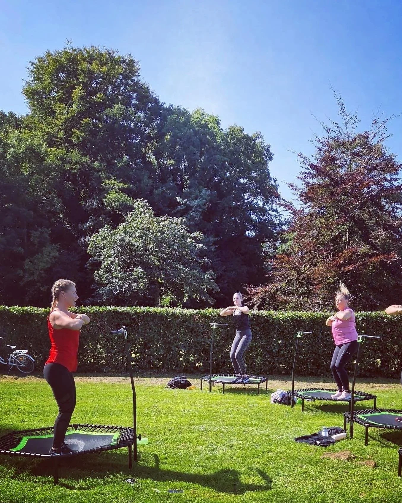 Tre kvinder træner på jump fitness udenfor med grønne træer i baggrunden og en blå himmel.