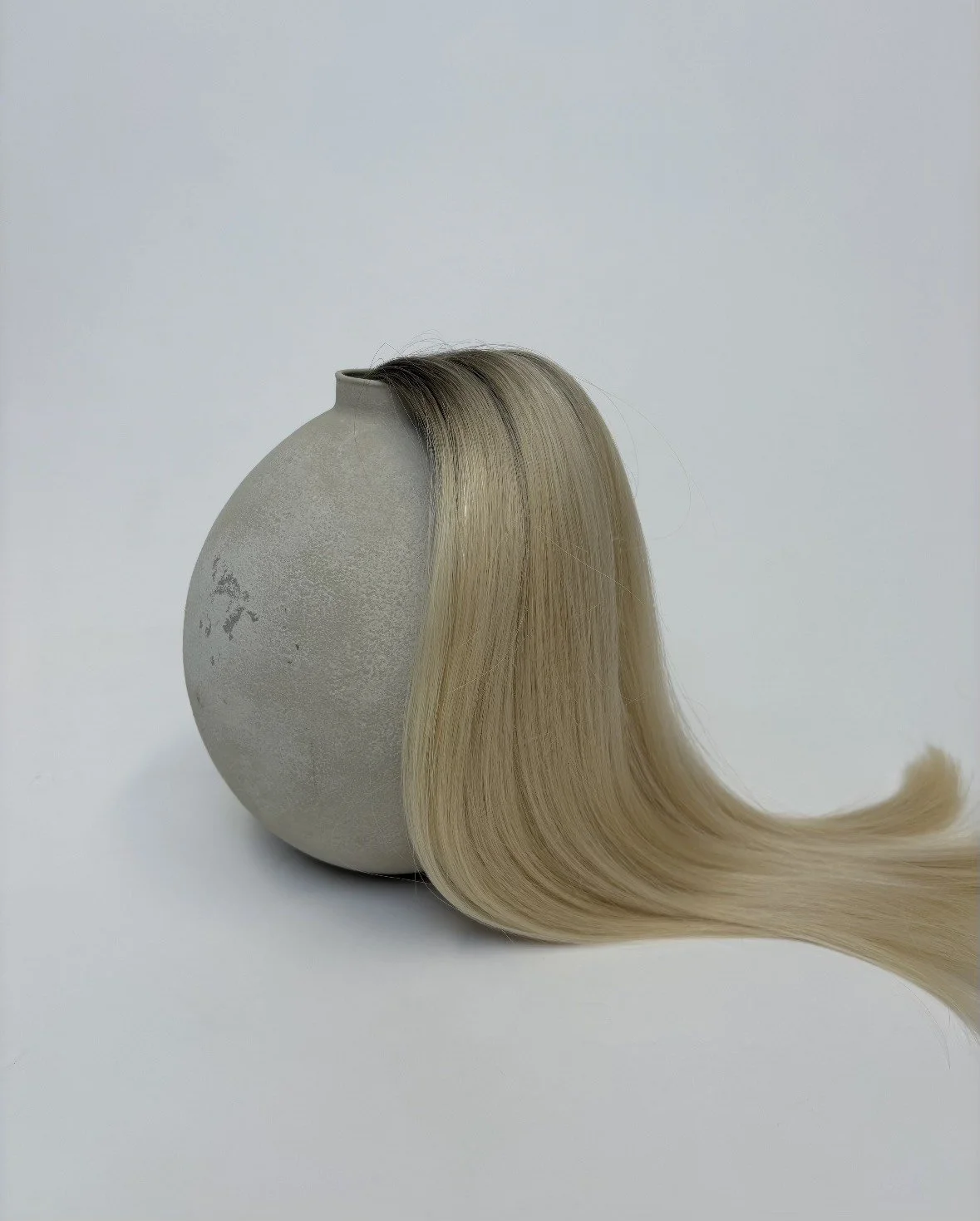 A round, textured, light gray ceramic vase with long, straight blonde hair flowing from its opening, set against a plain white background.