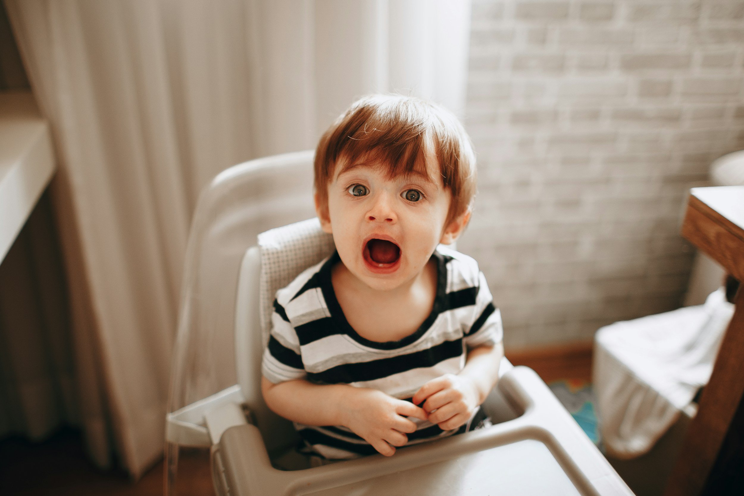 A young boy with brown hair and blue eyes sitting in a high chair, with his mouth open, appearing to be surprised or upset.
