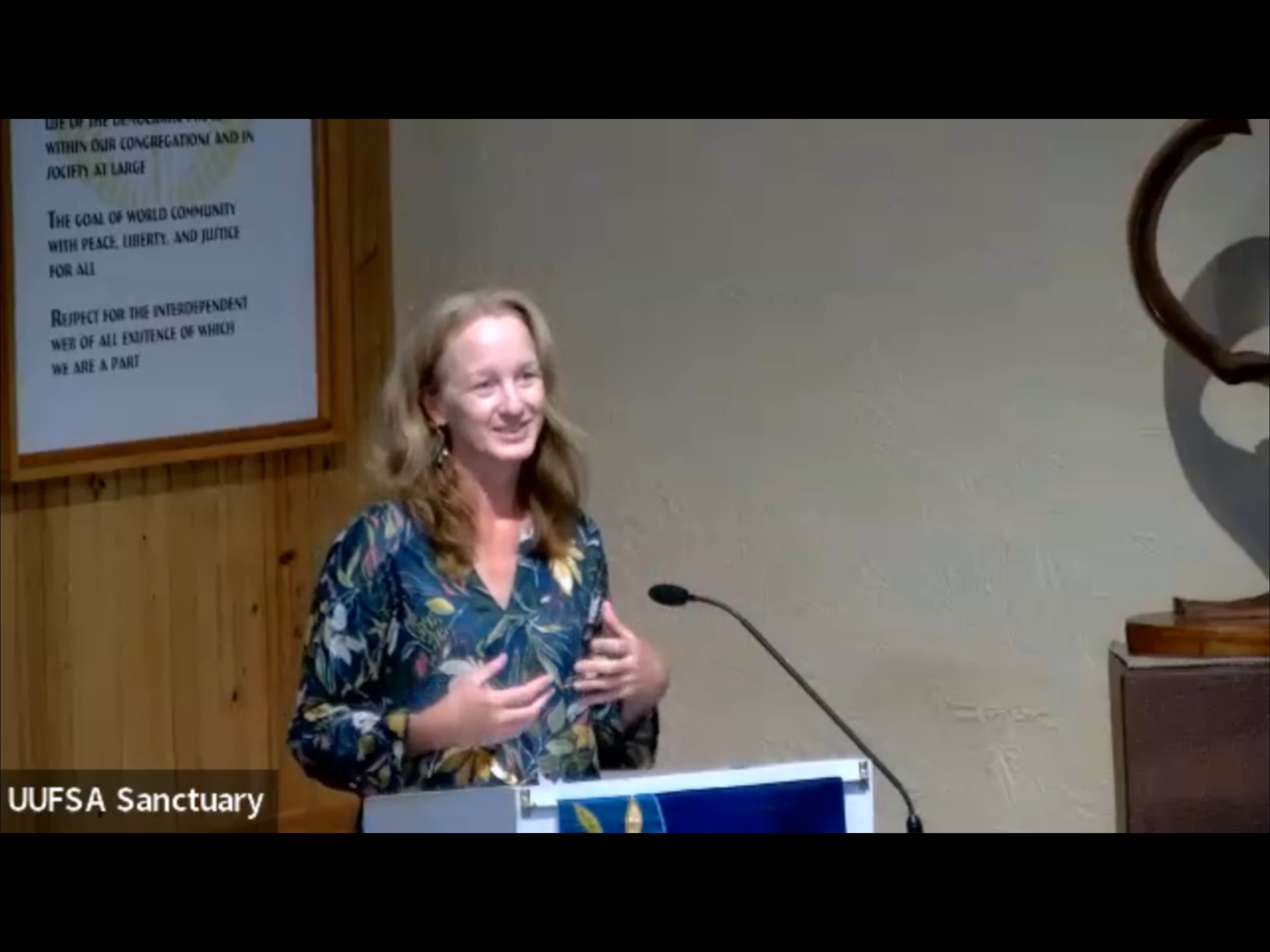 Woman smiling and speaking at a podium in a room with wooden paneling, a mural on the wall, and a sign that reads 'UUFSA Sanctuary'.