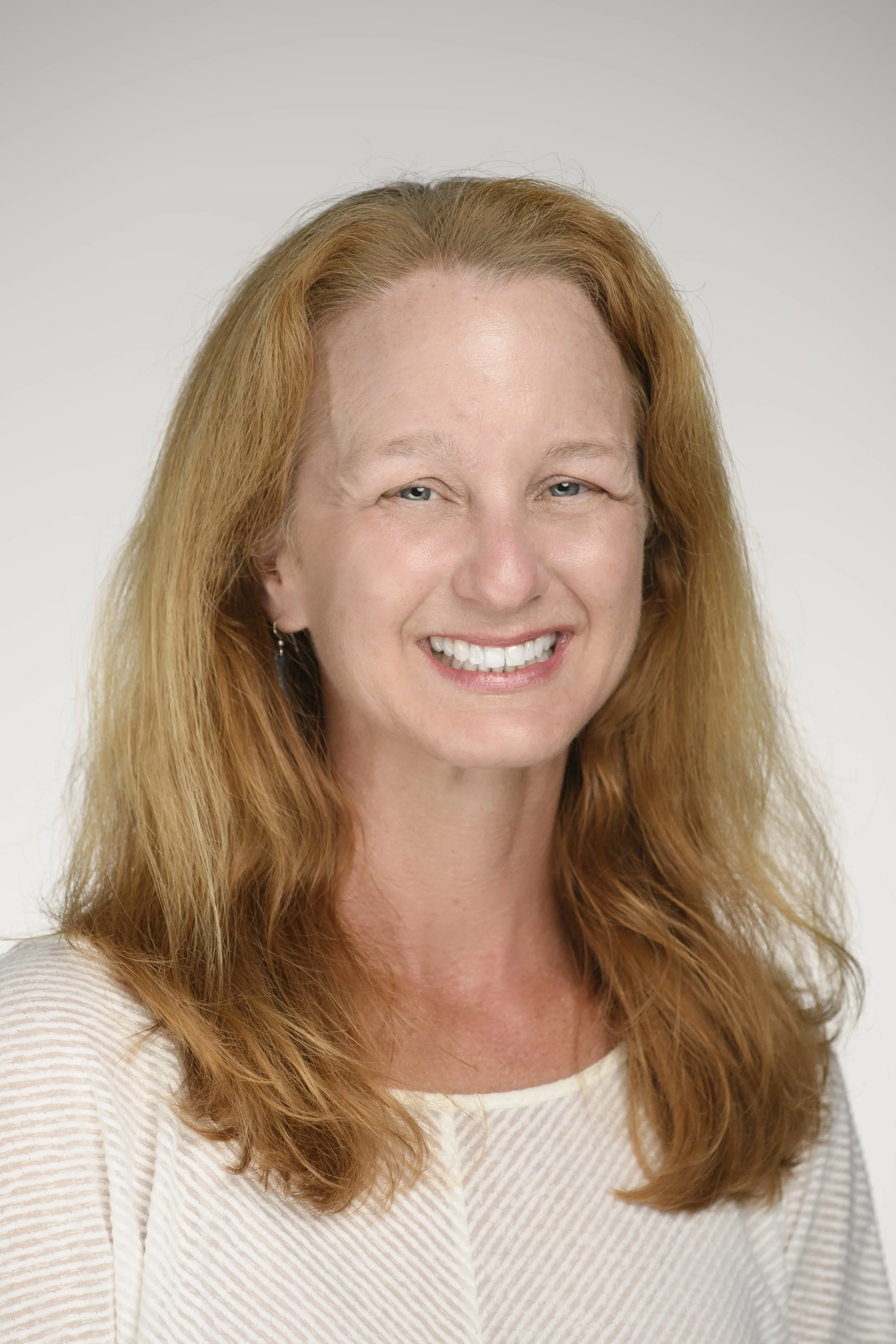 Portrait of a smiling woman with red hair, wearing a light-colored top, against a plain white background.