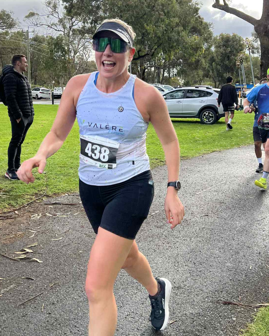 A woman running in a race, wearing a white tank top, black shorts, sunglasses, a black cap, and a race bib numbered 438. She appears to be happy and smiling.