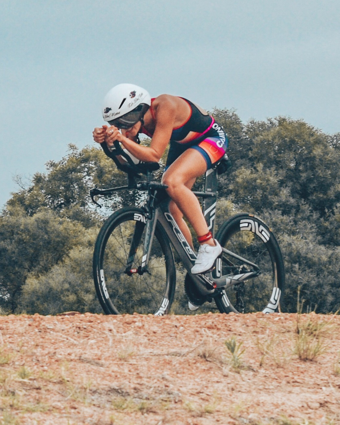 A triathlete wearing a helmet, sunglasses, and athletic gear riding a black aerodynamic bike on a dirt trail with trees and blue sky in the background.