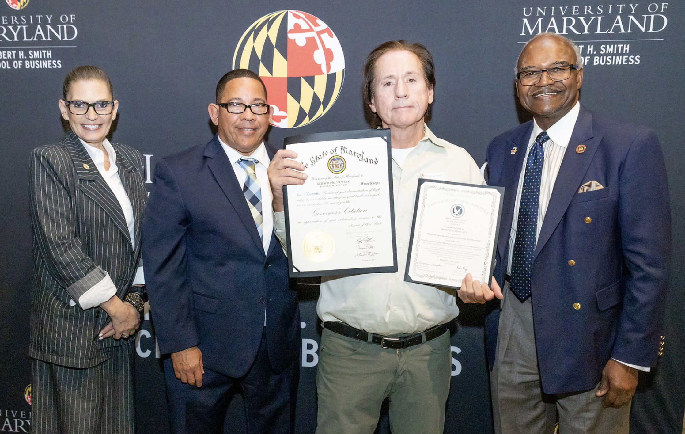 Group of five people, three men and two women, standing in front of a Maryland-related backdrop, holding certificates and smiling for a photo.