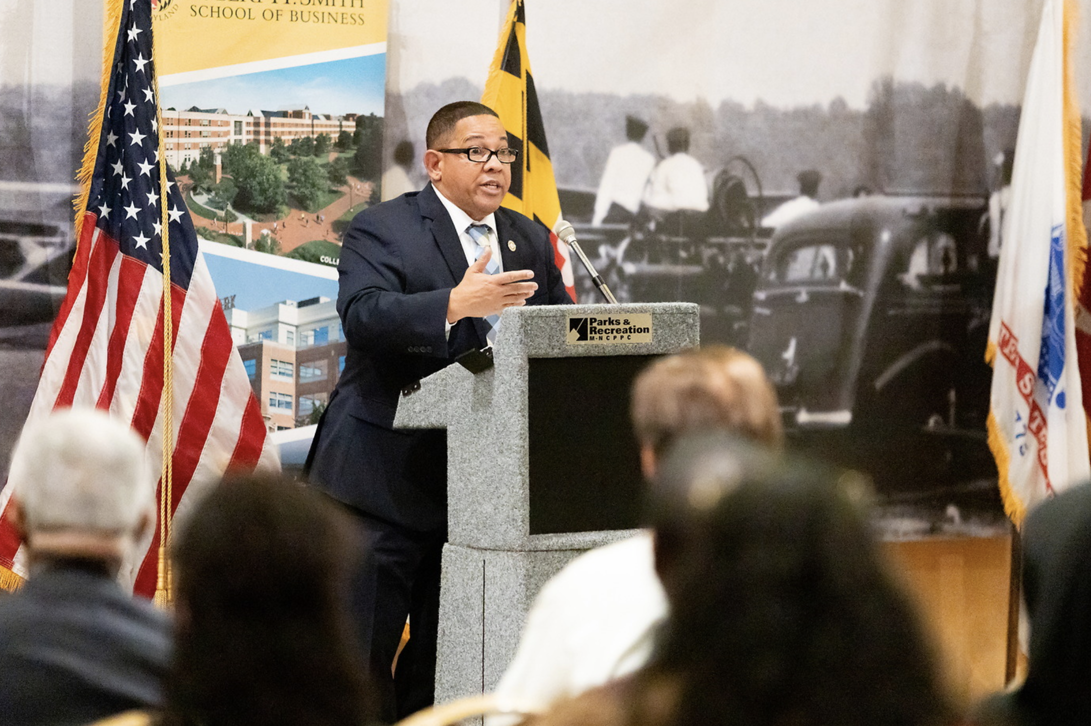 A man in a suit and glasses speaking at a podium with a microphone, in front of American and Maryland state flags, during an event at a school of business with a historical photo backdrop.