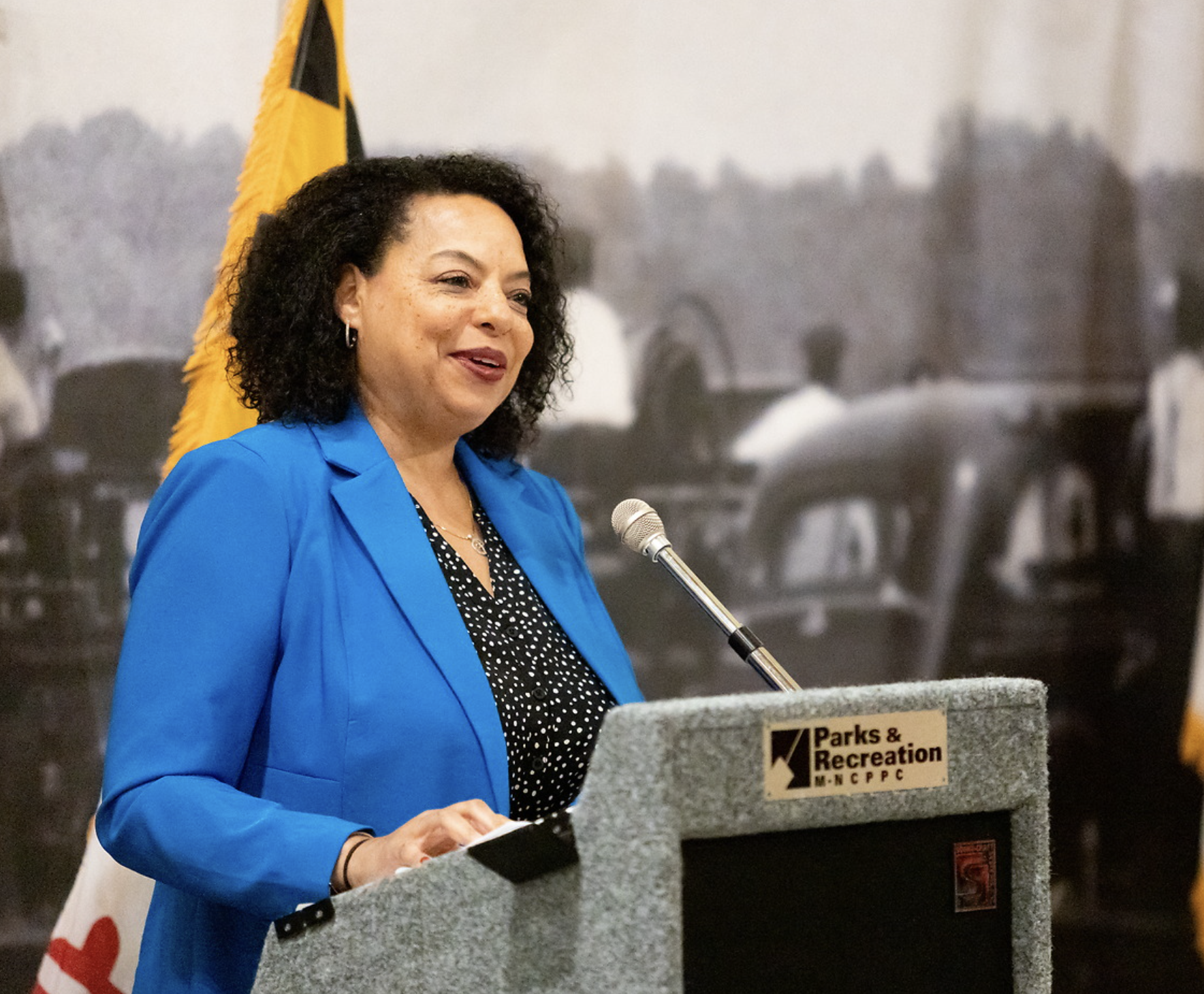 A woman with curly dark hair wearing a blue blazer and black- and-white polka dot shirt standing at a podium with a microphone, speaking at an event. There is a yellow and black flag behind her and a banner with the logo and text for Parks & Recreati