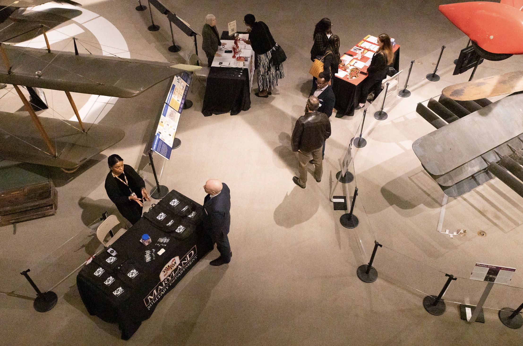 Museum exhibit with vintage gliders or small aircrafts on display, informational signage, and a registration table with brochures and memorabilia, with visitors exploring and engaging at the exhibit.