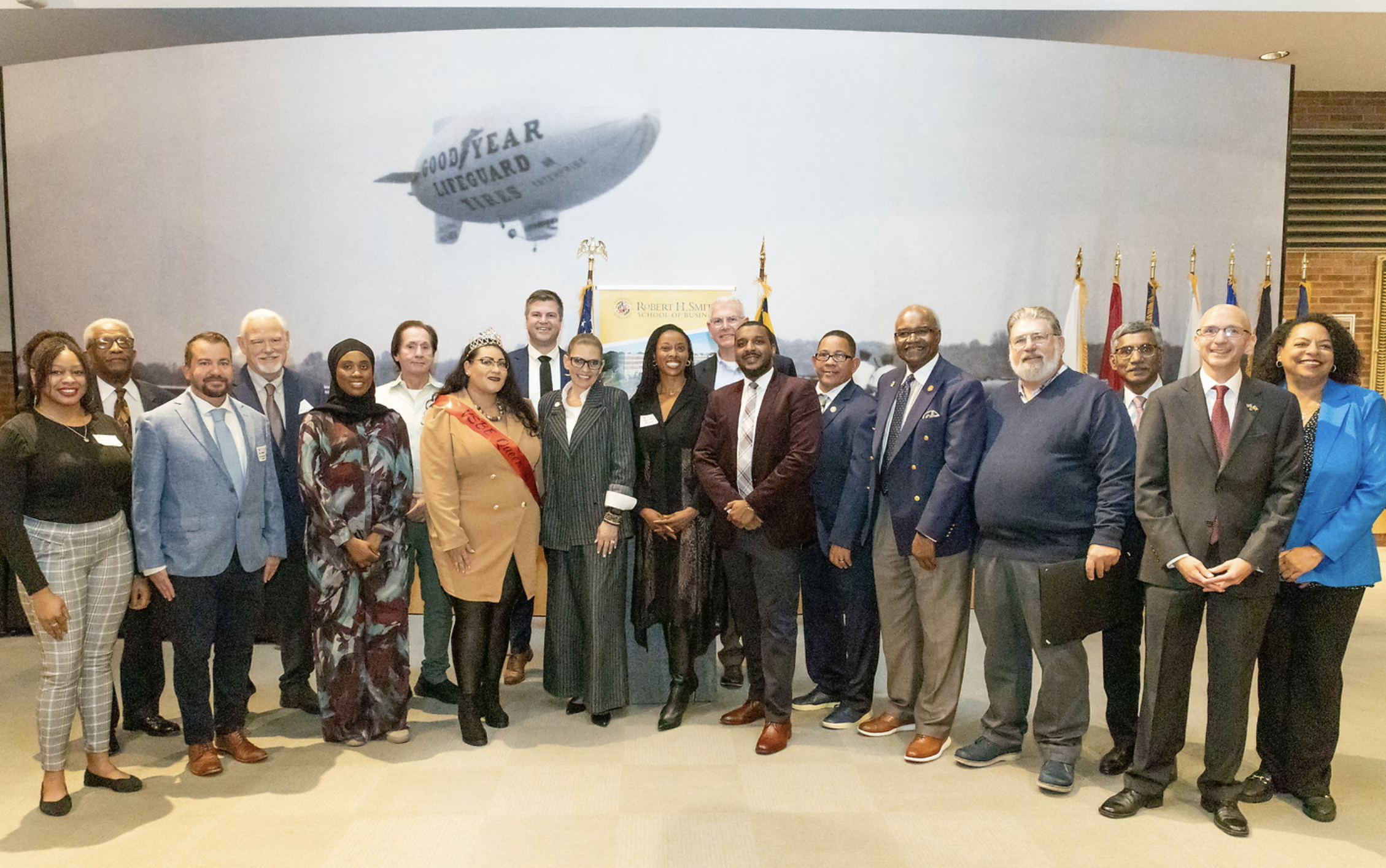 A group of diverse professionals posing together indoors at a formal event, with a backdrop of flags and a projected image of a blimp with Goodyear tires branding.