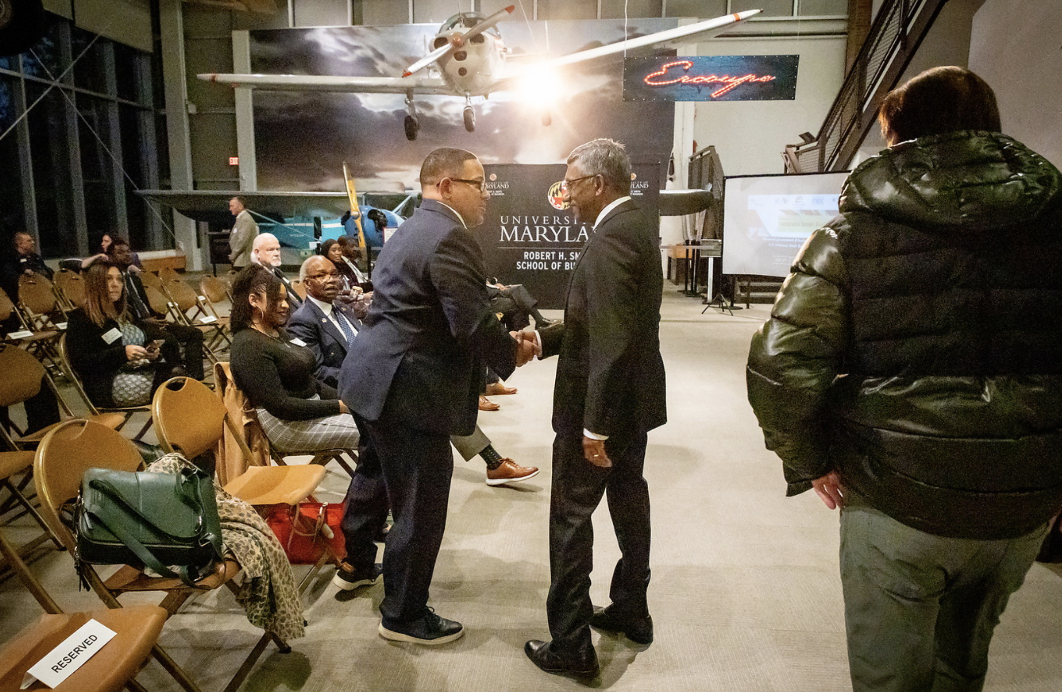 Two men in suits shaking hands in a conference room with seated attendees, aviation models, and a large aircraft image on the back wall.