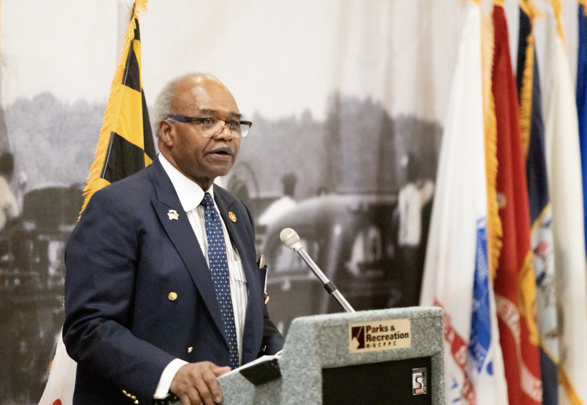 An elderly man in a dark suit with a white shirt and tie speaking at a podium, with American and other flags in the background, at a Parks & Recreation event.