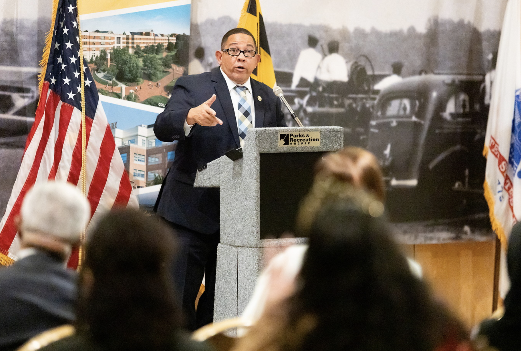 A man in a suit and glasses speaking at a podium with a "Parks & Recreation" sign, flanked by American and Maryland flags, with a backdrop of historical photos and a colorful illustration of a park or campus.