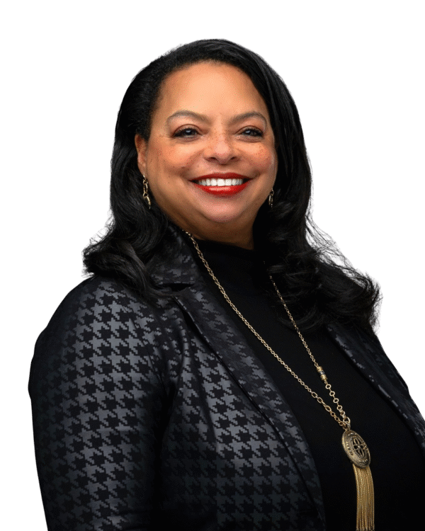 A smiling African American woman with dark hair, wearing a black and gray houndstooth-patterned blazer, gold earrings, and a long gold necklace with a circular pendant, against a black background.