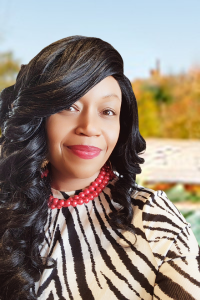 Portrait of a woman with long black hair and a patterned dress, outdoors with trees and a building in the background.