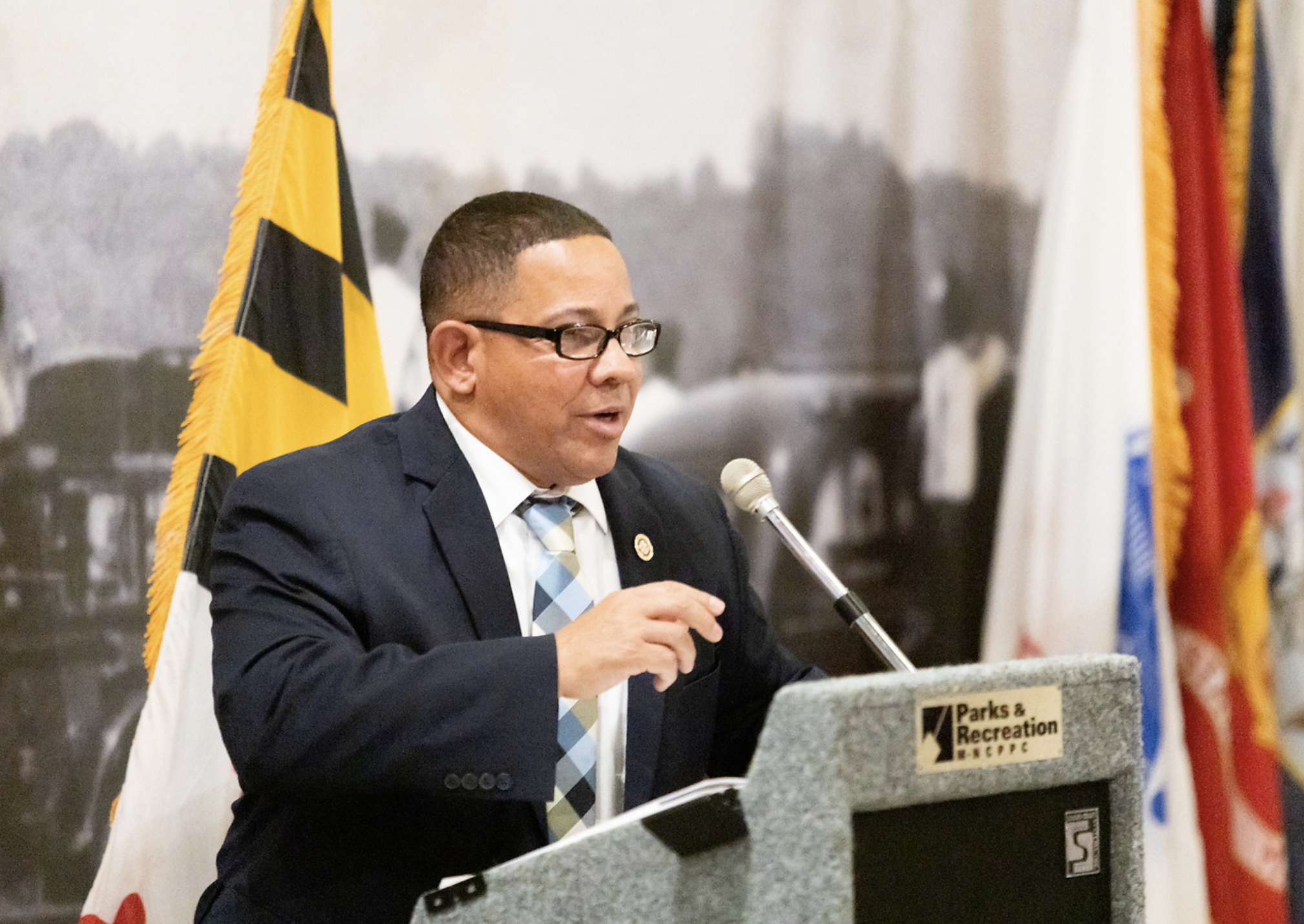 A man in a suit and glasses speaking into a microphone at a podium in front of flags, with a sign that reads 'Parks & Recreation'}