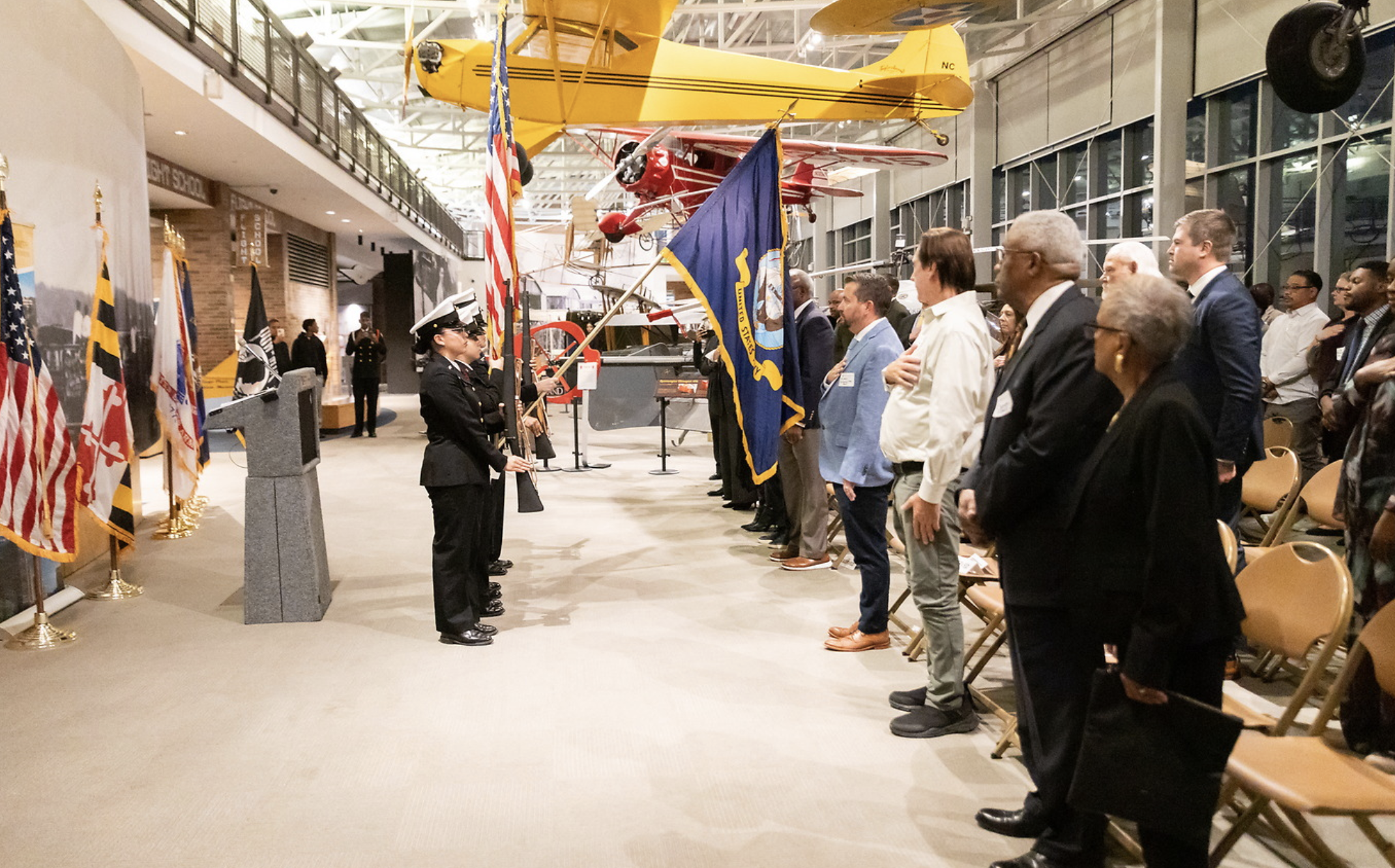 A group of people standing in a line with their right hands over their hearts during a ceremony at an indoor aviation museum. In the background, there are two vintage airplanes hanging from the ceiling, a yellow one, and a red and white one. Several 