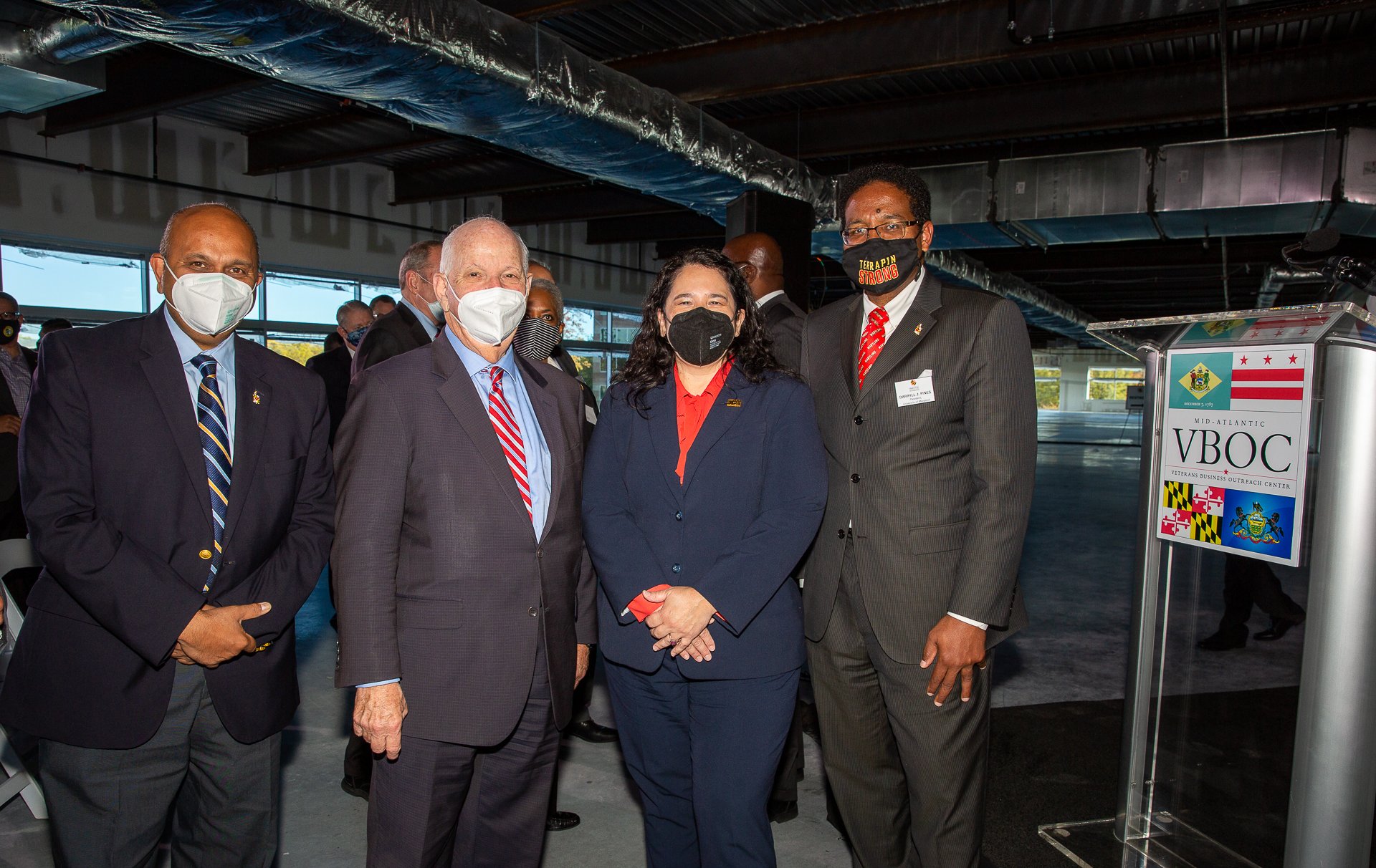 Four individuals wearing suits and face masks standing together at an indoor event, with a sign pointing to the Veterans Business Outreach Center in the background.