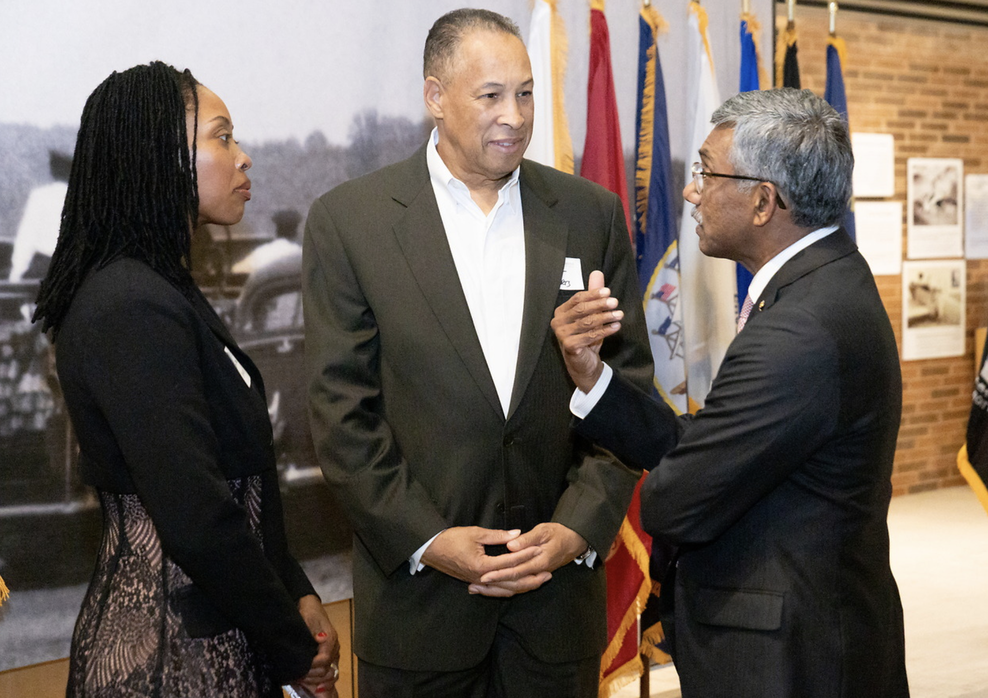 Three individuals engaged in conversation at an indoor event. A woman with long black dreadlocks wearing a black blazer, a man with short hair wearing a dark suit, and an older man with gray hair and glasses. Background features flags and historical 