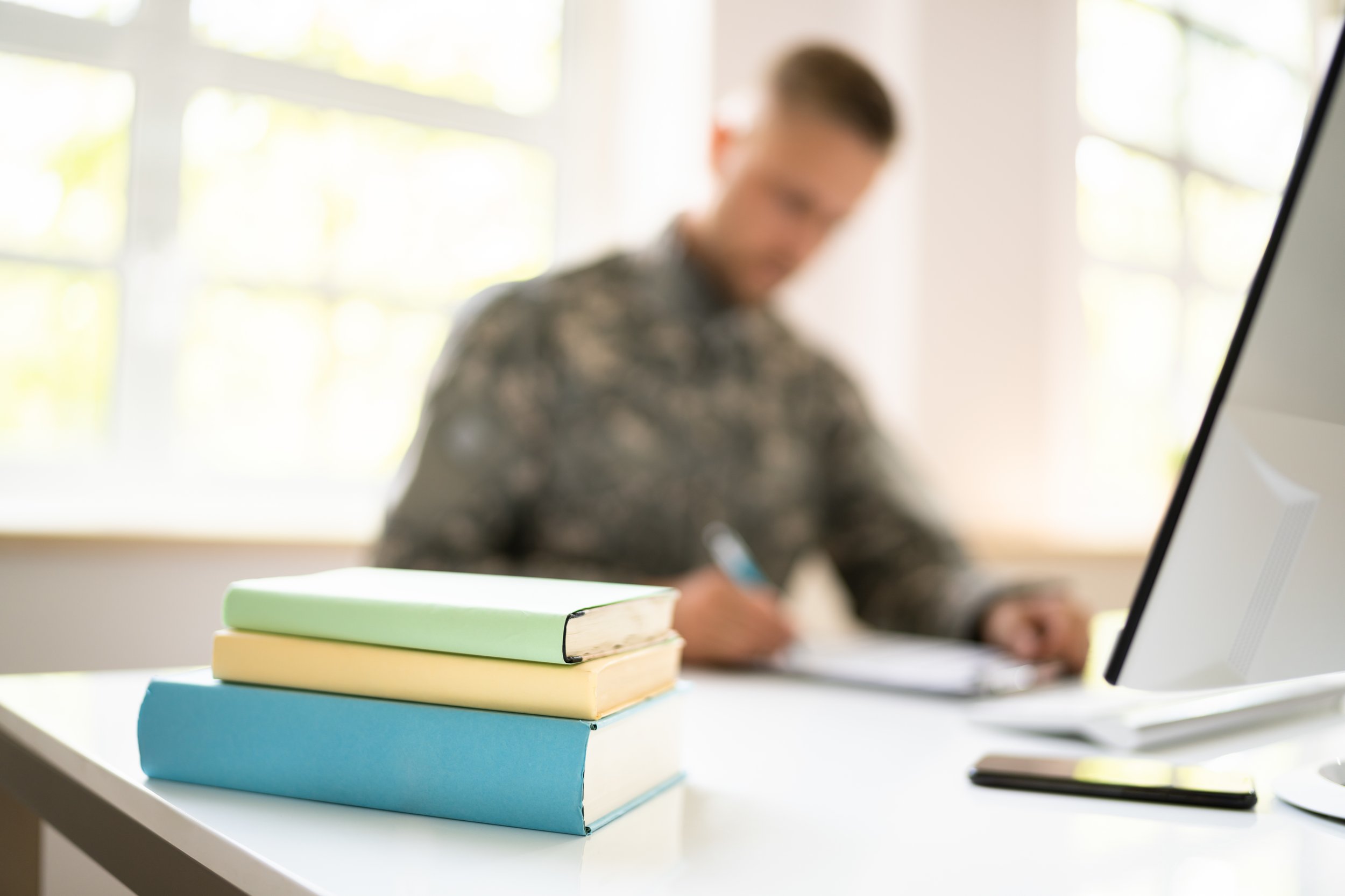 A white desk with three stacked hardcover books in pastel green, yellow, and blue in the foreground, and a person working at a computer in the background.