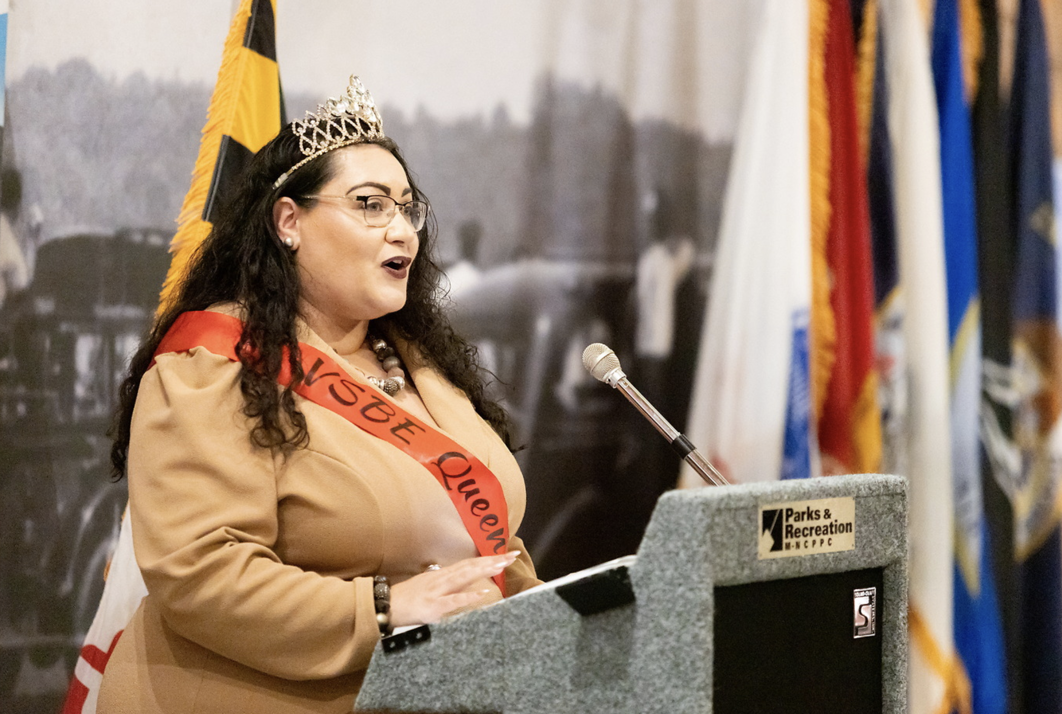A woman wearing glasses, a tiara, a beige top, and a red sash stands at a podium speaking into a microphone, with flags in the background.