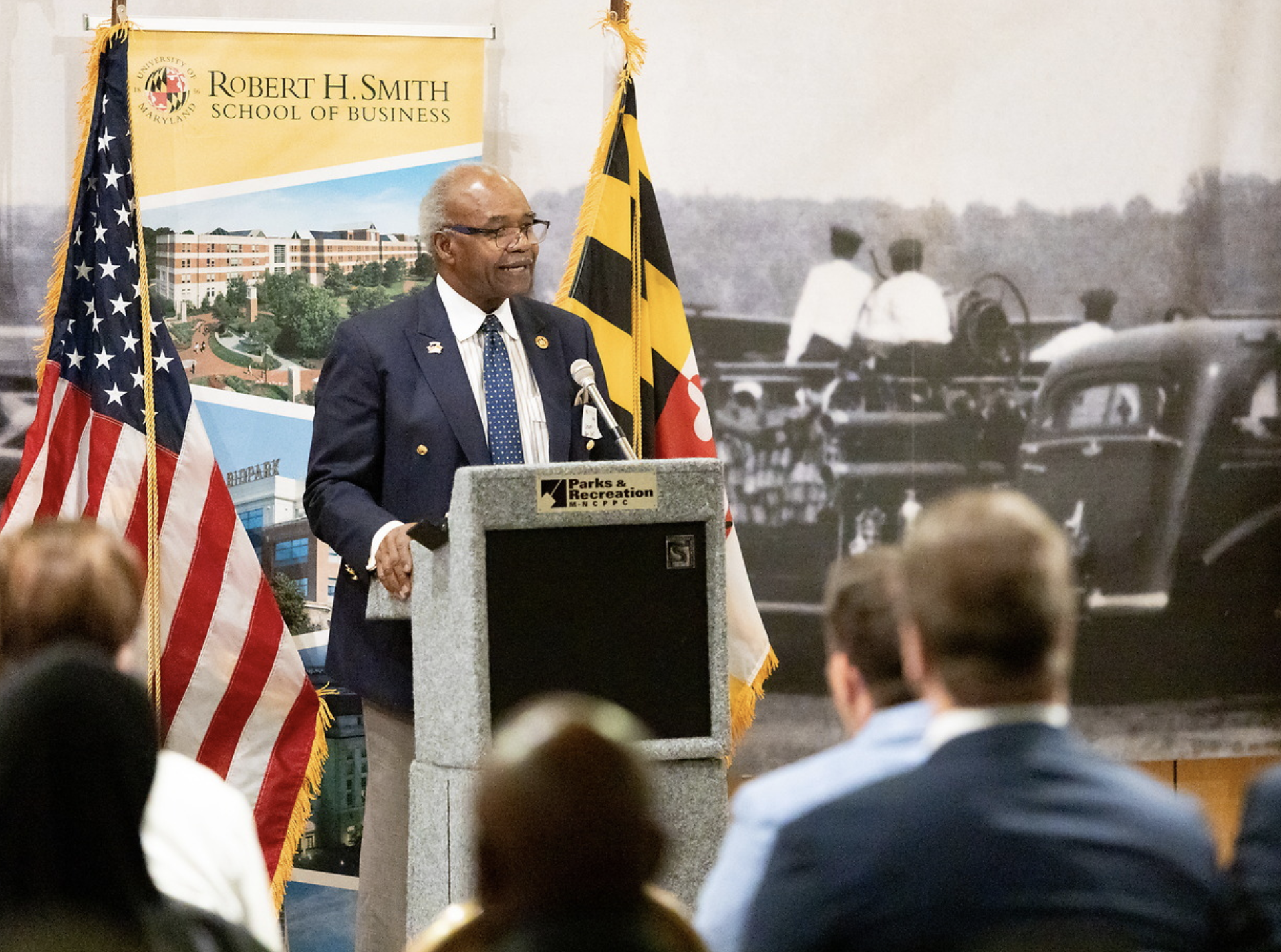 An older man with white hair and glasses giving a speech at a podium with the Parks & Recreation Department logo, flanked by the American flag and a Maryland state flag, in front of a large backdrop featuring a historic black-and-white photo of child