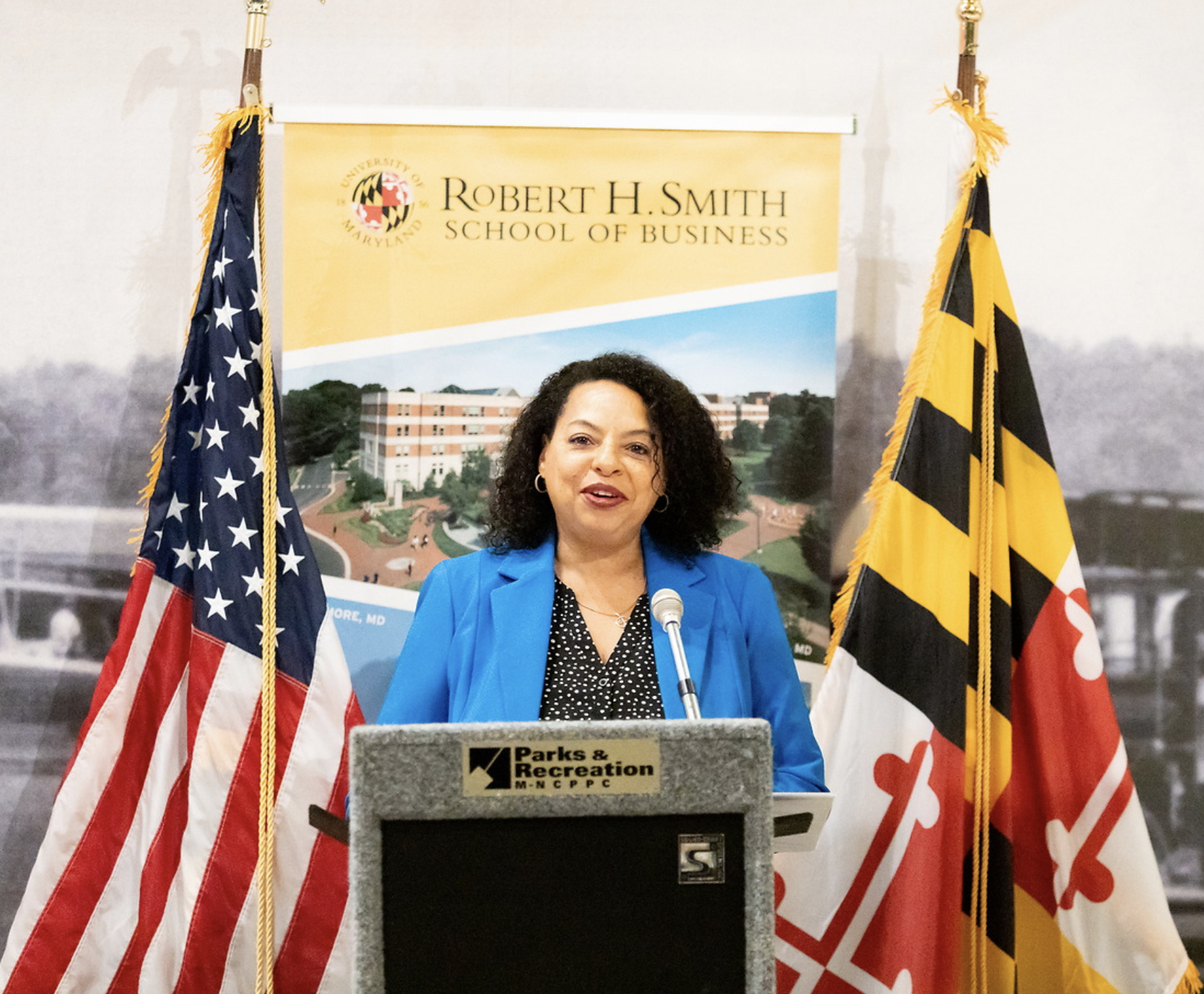 A woman in a blue blazer speaking at a podium with a microphone, with American and Maryland state flags behind her. A banner for the Robert H. Smith School of Business is in the background.
