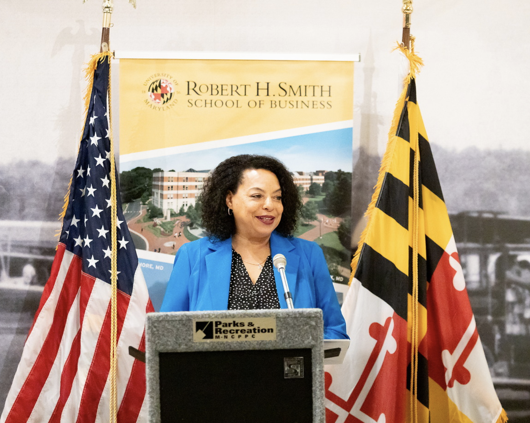 A woman with curly black hair and wearing a blue blazer and black polka dot blouse stands at a podium, speaking into a microphone. Behind her are two flags—one American and one Maryland—and a yellow banner with the text 'Robert H. Smith School of Bus