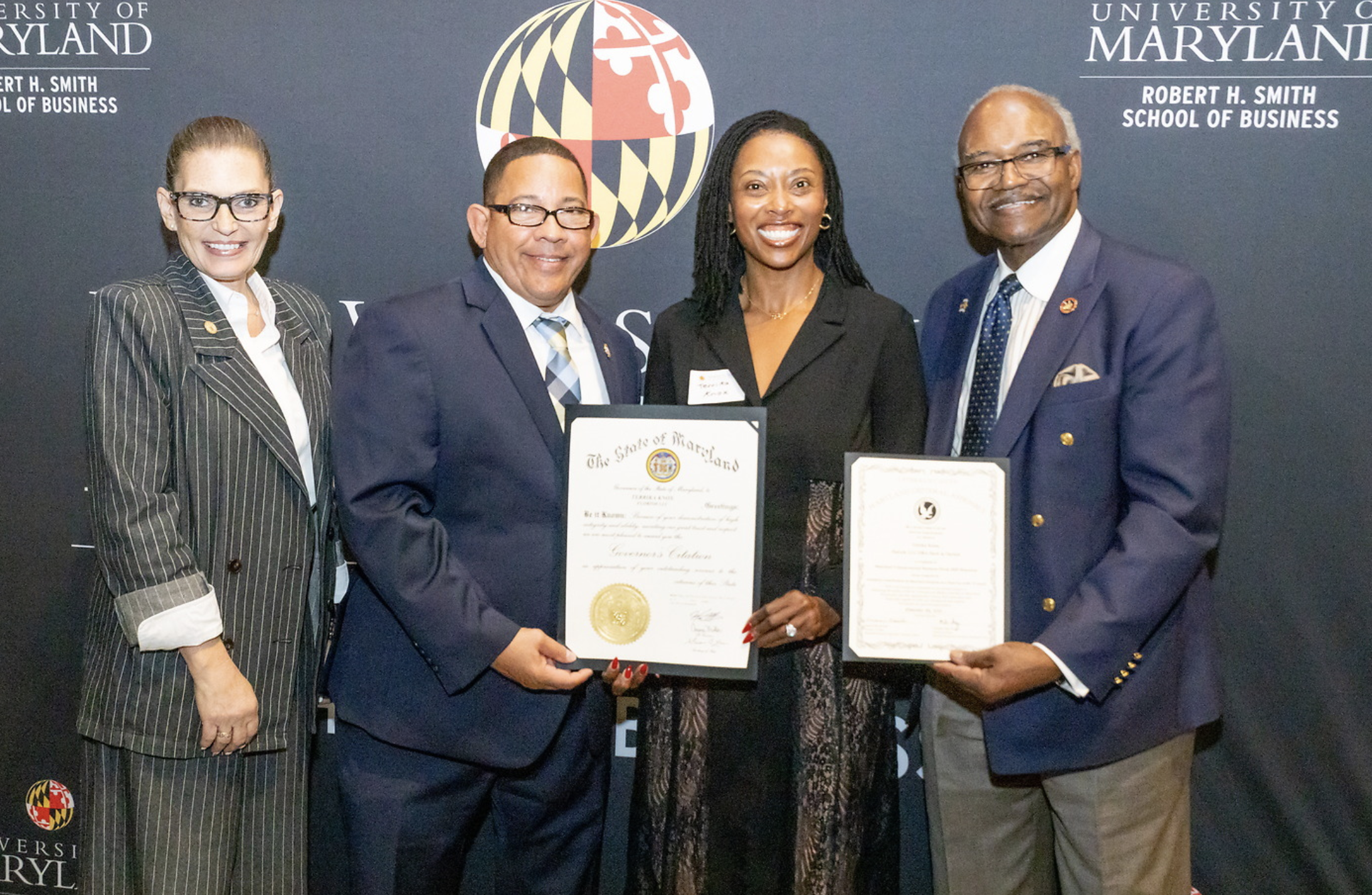 Four individuals in professional attire standing together, holding certificates, in front of a backdrop with the University of Maryland logo and text.