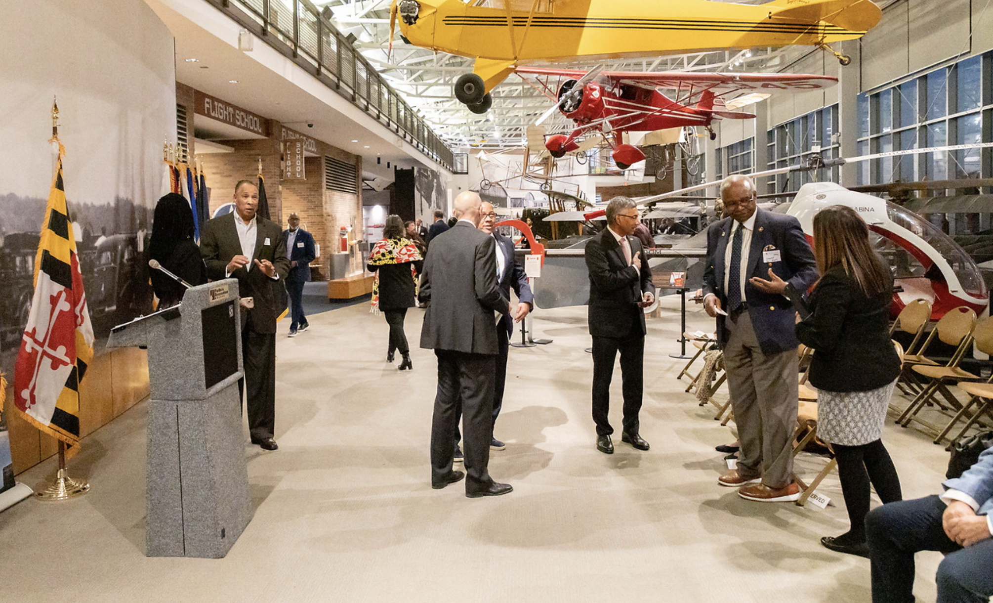 People gathered in an indoor exhibition space with vintage aircraft on display overhead. A speaker at a podium is addressing an audience, with some individuals engaged in conversation and others seated. Flags and informational signs are visible, indi