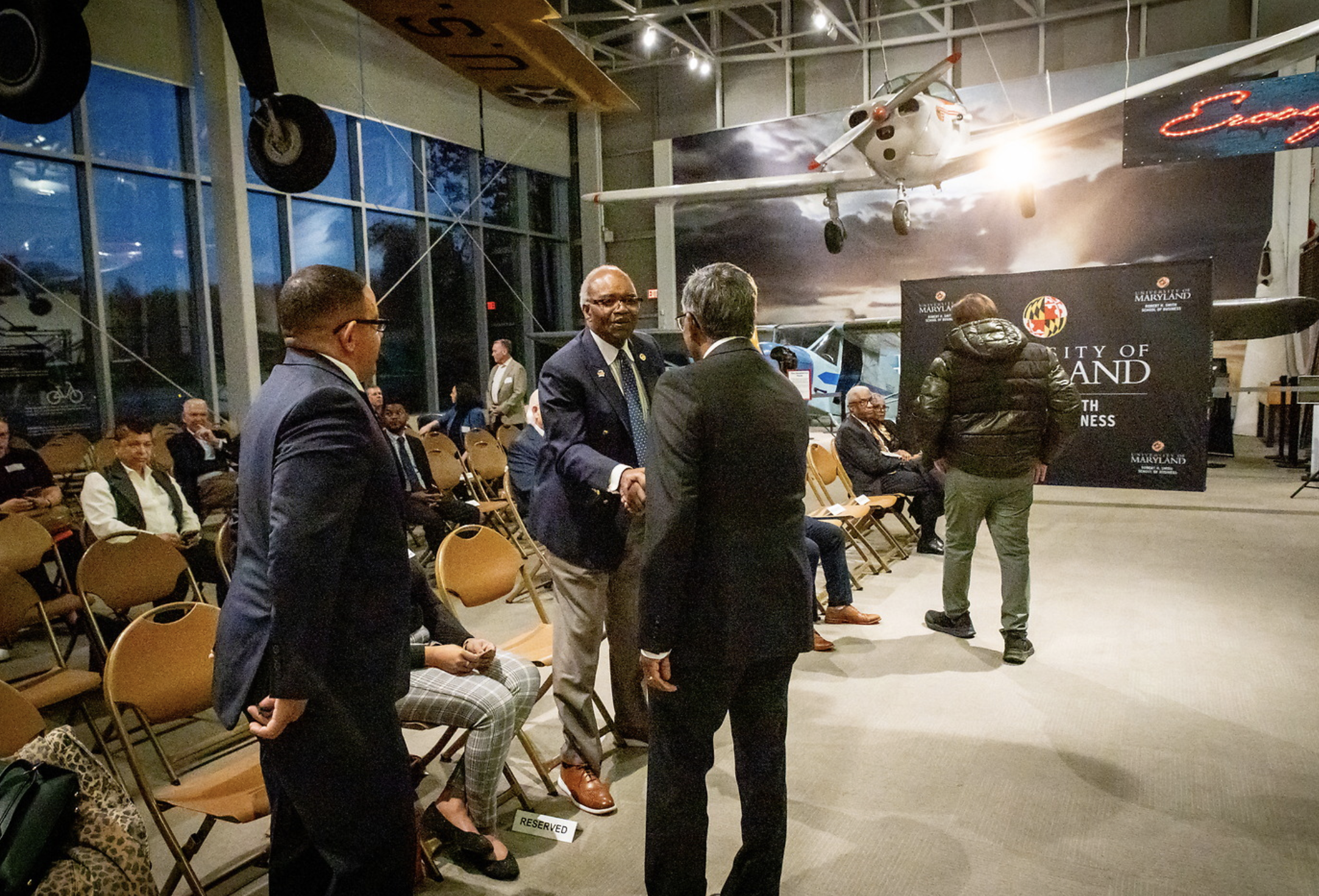 A group of people in suits and business attire are gathered in a large indoor space with high ceilings and glass walls. Some are engaged in conversation, while others are sitting on chairs. In the background, there are aircraft models, including a sm