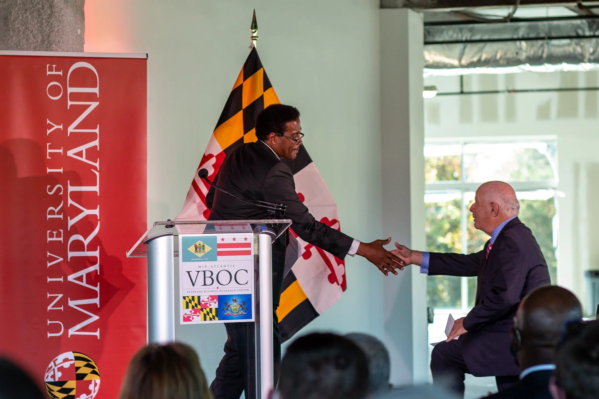 Two men shaking hands on a stage at a formal event, with Maryland and Baltimore flags visible behind them. One man is standing at a podium with a Maryland flag and VBOC sign, while the other is seated. Audience members are visible in the foreground.