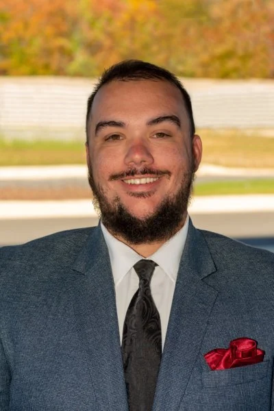 A smiling man with a beard and short hair, wearing a gray suit, white shirt, dark tie, and red pocket square, outdoors with trees and a fence in the background.