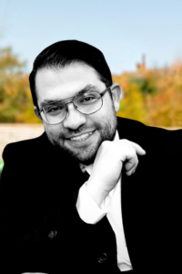 A man with glasses and dark hair smiling outdoors, with a blurred background of trees and sky.