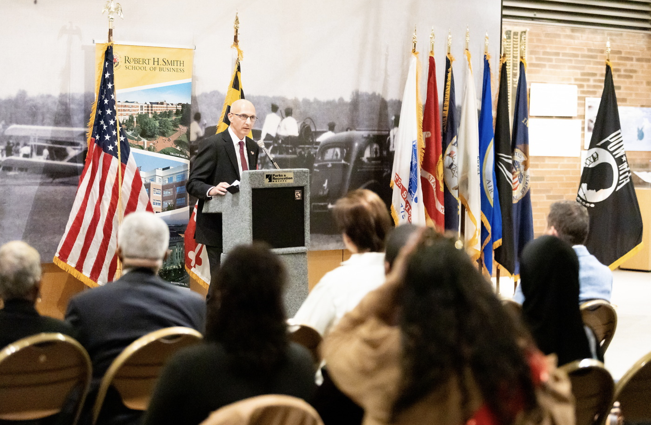 A man speaking at a podium during an event at Robert H. Smith School of Business, with multiple flags behind him and a large black-and-white historical photo on the wall.
