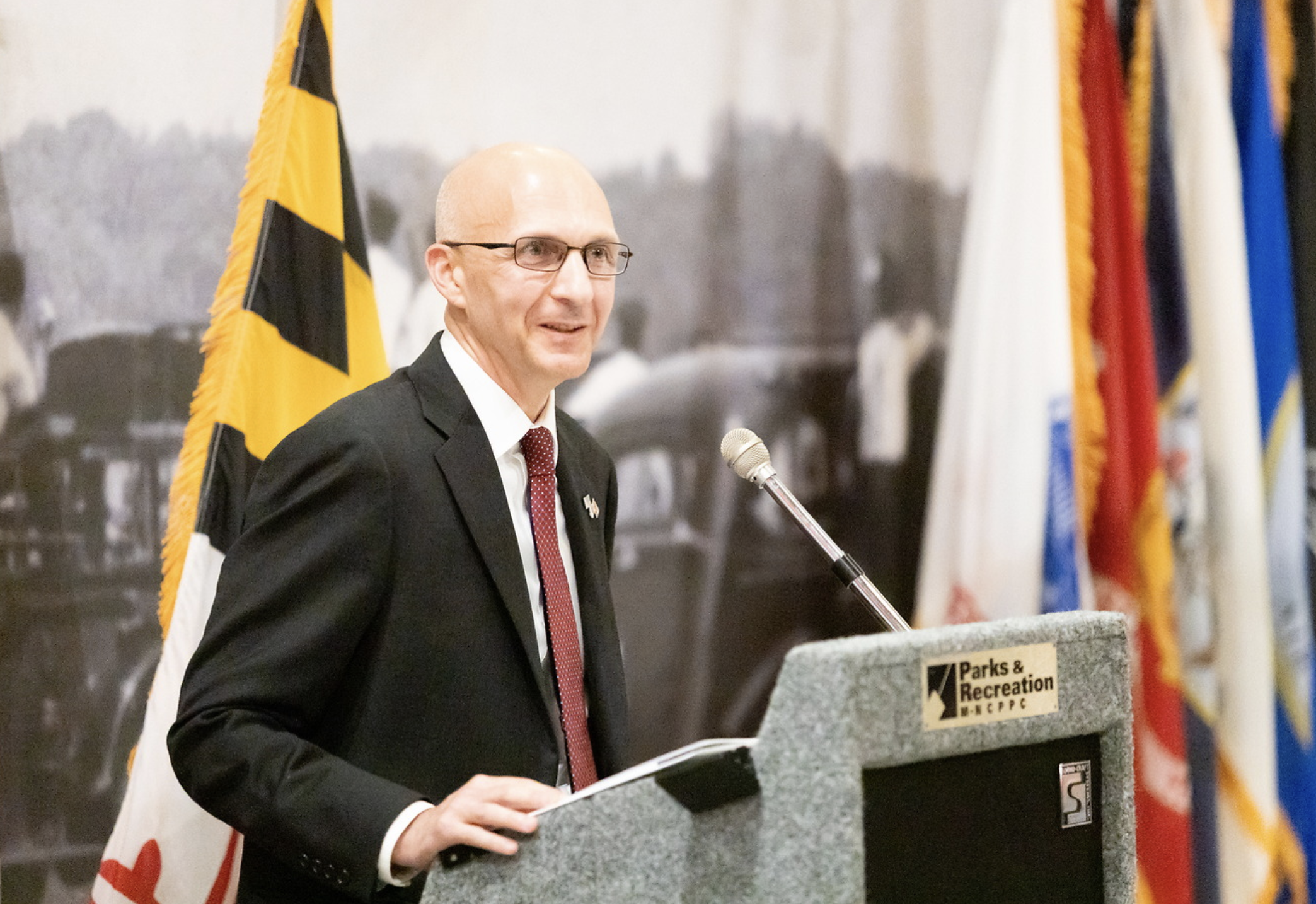 A man in a black suit and red tie speaking at a podium with flags in the background.