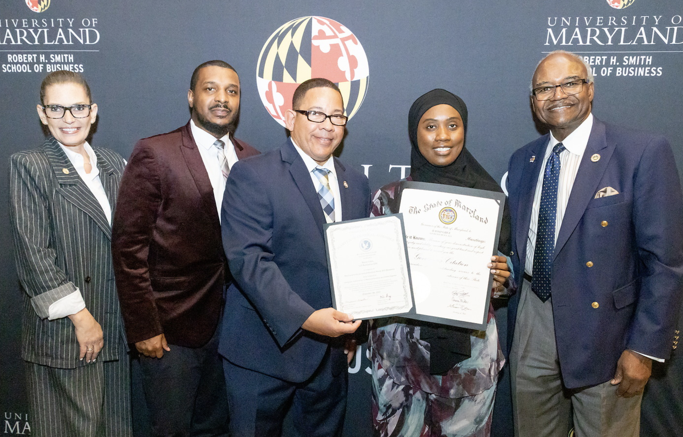 Group of five diverse people dressed in formal attire, holding framed certificates, standing in front of a dark blue UNM and University of Maryland backdrop at a formal event.