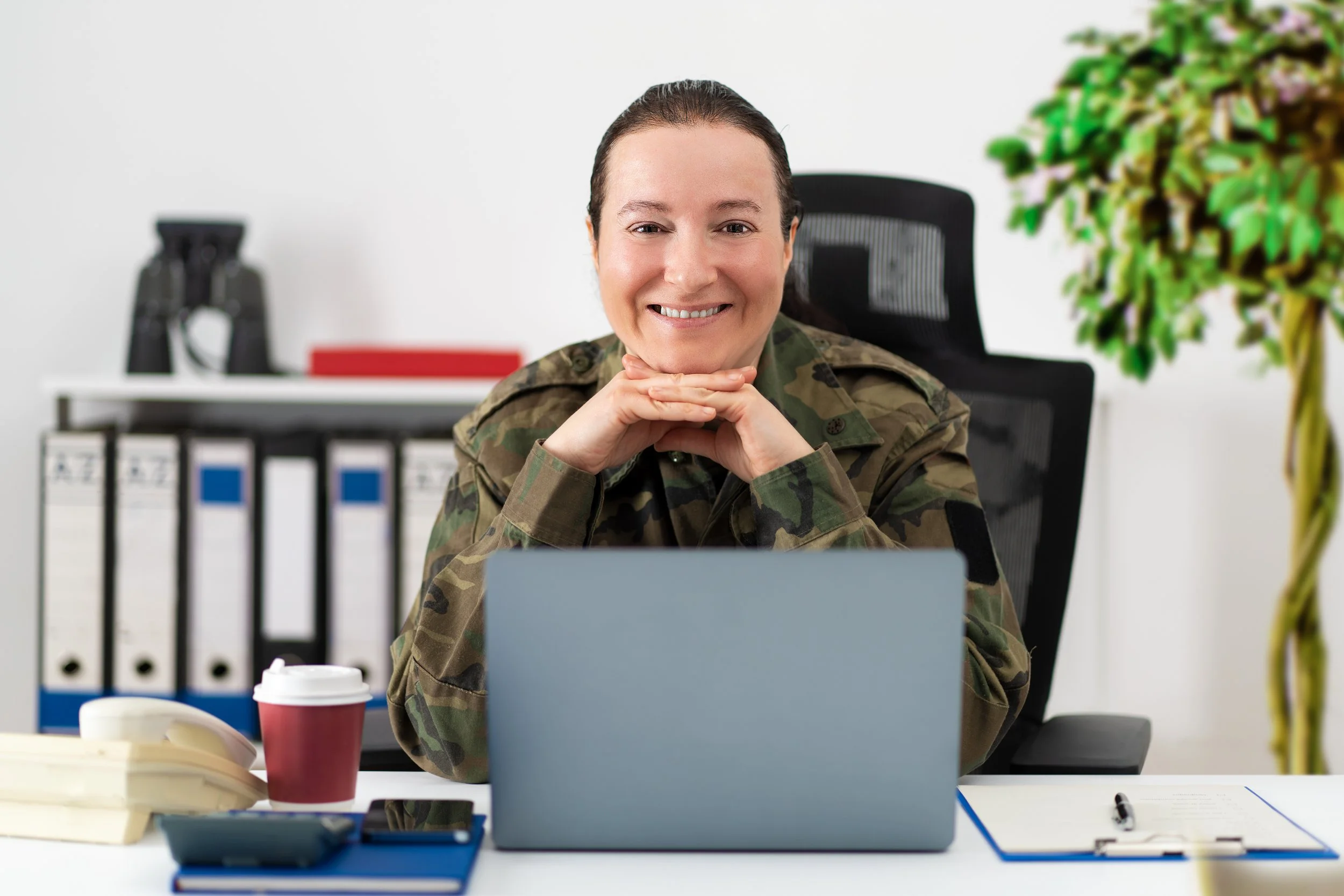 Woman in military uniform smiling at camera, sitting at an office desk with a laptop, coffee cup, phone, and files.