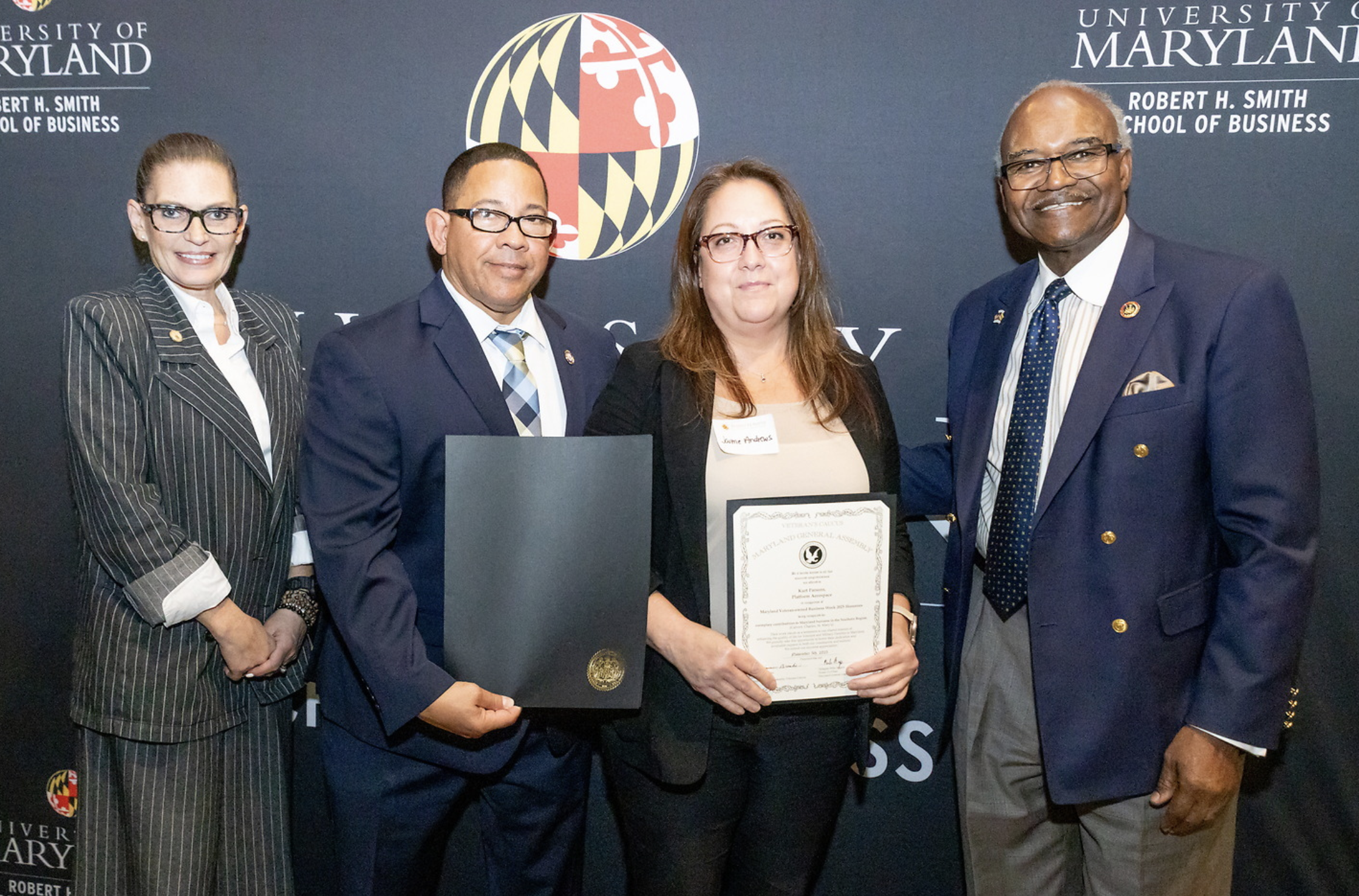 Group of four people standing in front of a University of Maryland backdrop. One woman in the center is holding a certificate and a folder, and all are dressed in business attire.
