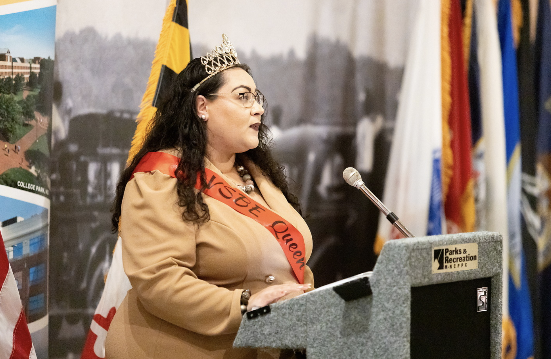 A woman wearing glasses, a crown, and a sash is speaking at a podium. She is dressed in beige and speaking into a microphone. Behind her are several flags and a backdrop with cityscape images.